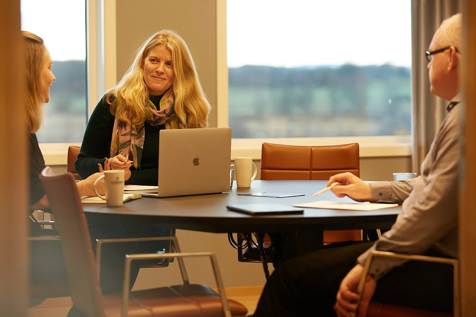 Three people sitting around a conference table in a meeting room with a large window showing an outdoor landscape, engaged in conversation. The woman in the center has blonde hair, is wearing a colorful scarf, and is working on a laptop. The other woman on the left and the man on the right are listening.