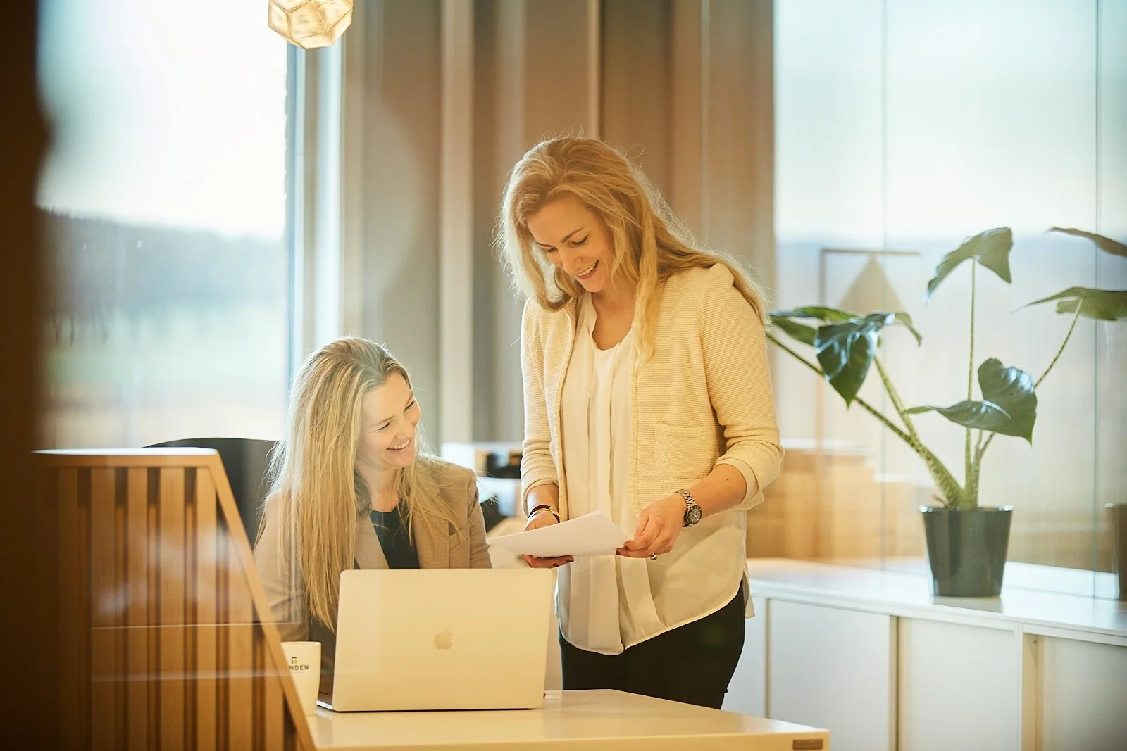 Two women are in a bright office, one sitting at a desk working on a laptop and the other standing, holding papers, smiling and engaging in conversation.
