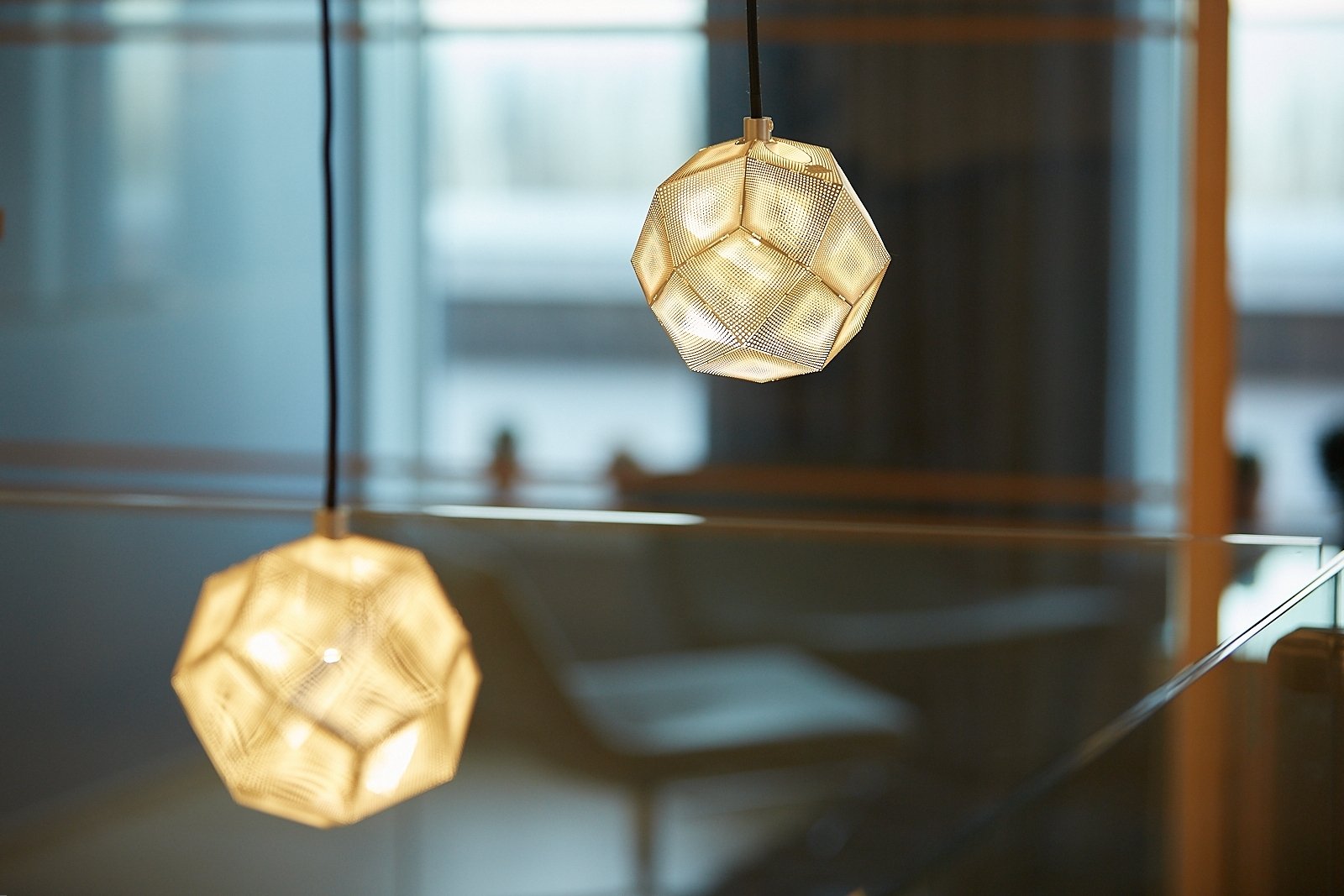 Two modern pendant lights with geometric, multifaceted gold-colored shades hanging from the ceiling inside a room with large windows.