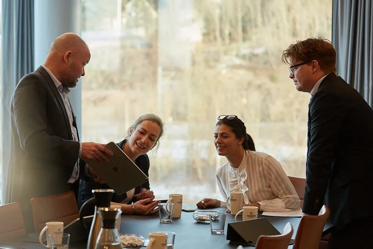 Four professionals in a meeting room with a large window, two men and two women, engaged in a discussion, with coffee mugs, glasses, and a laptop on the table.