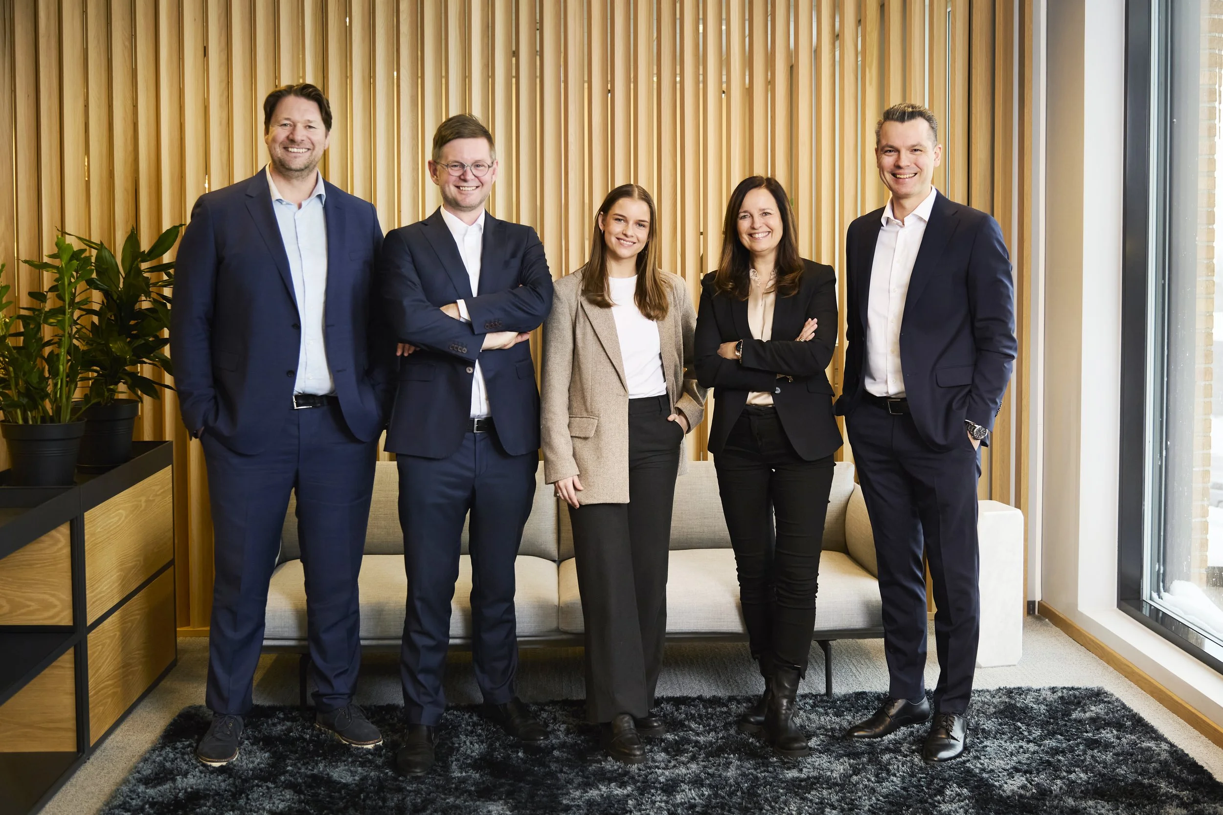 Group of five professionals in business attire standing in a modern office with wood panel walls and a window.