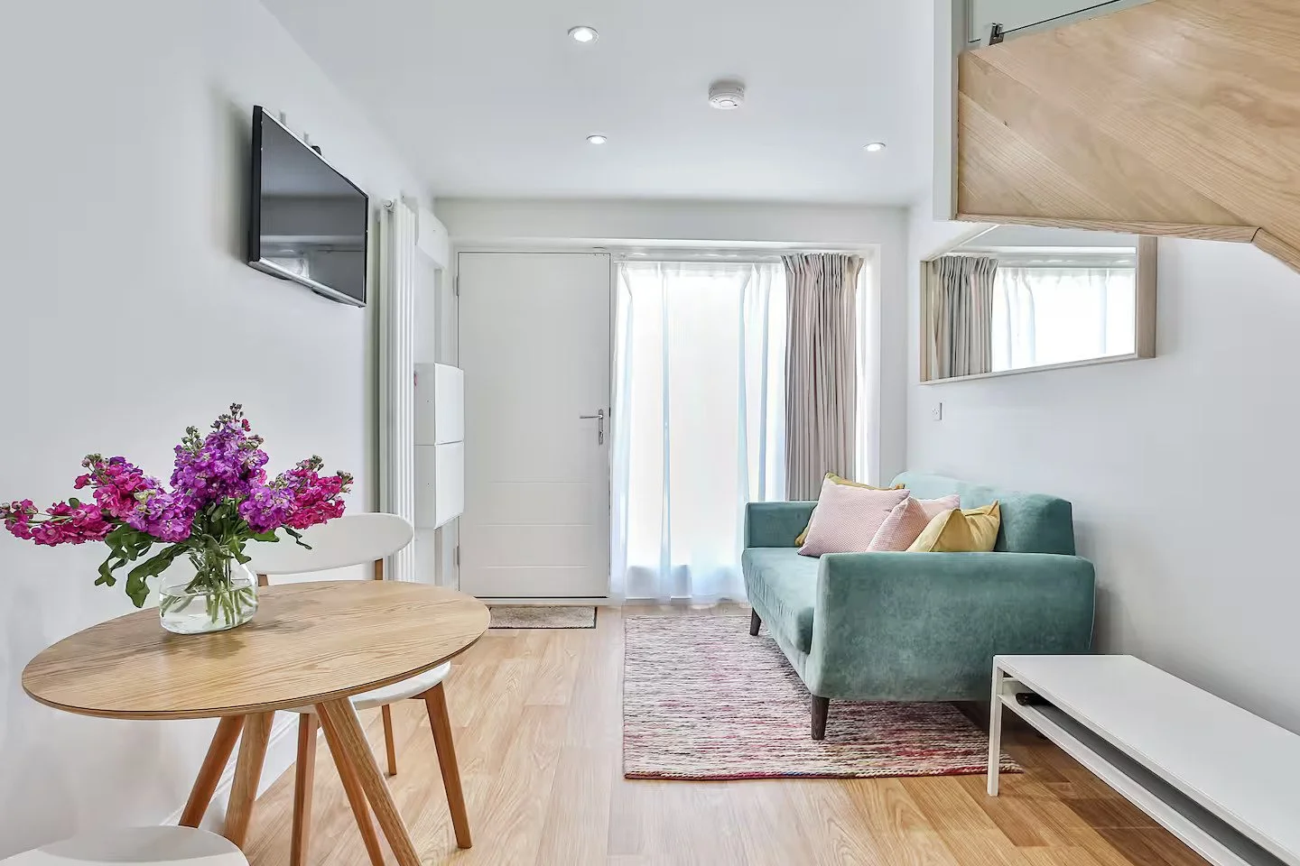 Living room with light-colored walls, a mounted flat-screen TV, a small wooden dining table with a vase of purple and pink flowers, a green sofa with pastel cushions, a patterned rug, curtains, a wall mirror, and a white TV stand.