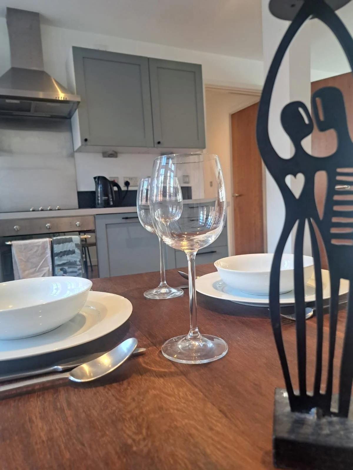 A dining table with three wine glasses, bowls, plates, and cutlery, set in a modern kitchen with gray cabinets and a stovetop.