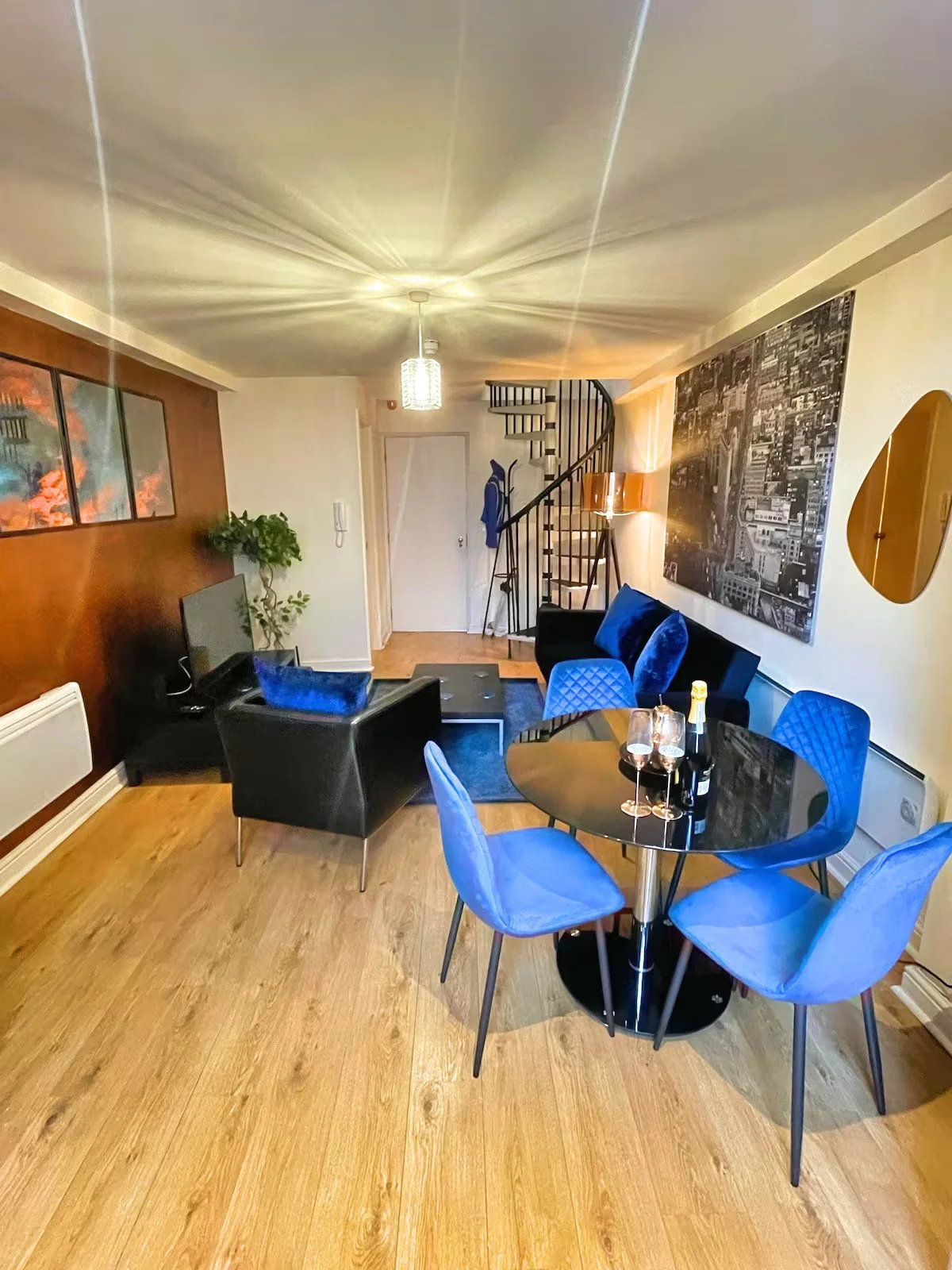 A living room with a black sofa, blue cushions, a black armchair, a round black dining table with blue chairs, a television on a stand, a plant, a spiral staircase, a large black-and-white cityscape photograph on the wall, and a bottle of champagne with two glasses on the table.