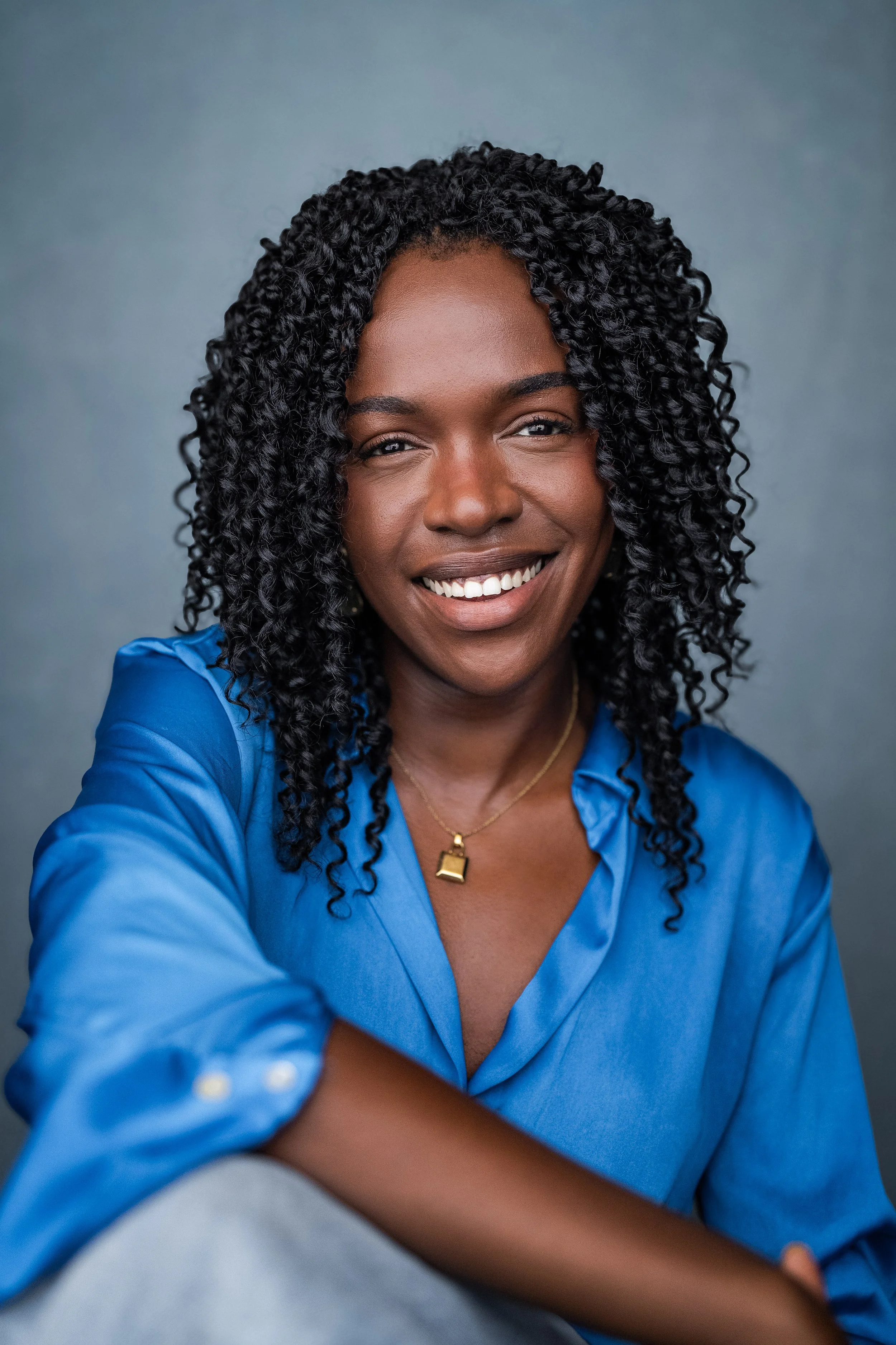 A smiling Black woman wearing a blue shirt and a gold necklace, against a plain gray background.