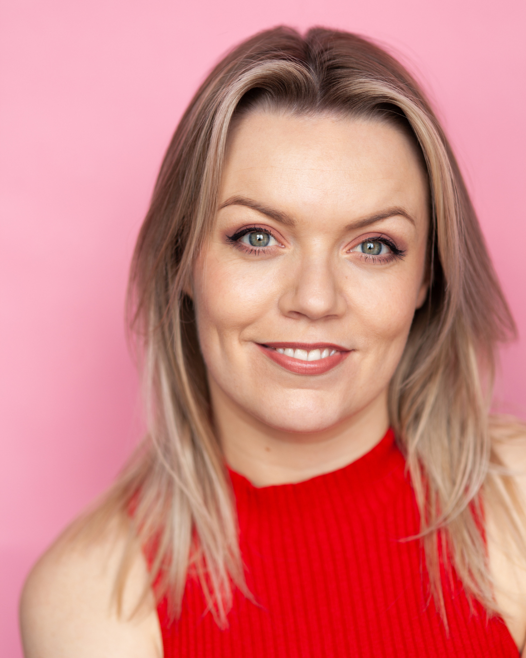A close-up portrait of a smiling woman with blonde hair and blue eyes, wearing a red sleeveless top, against a pink background.