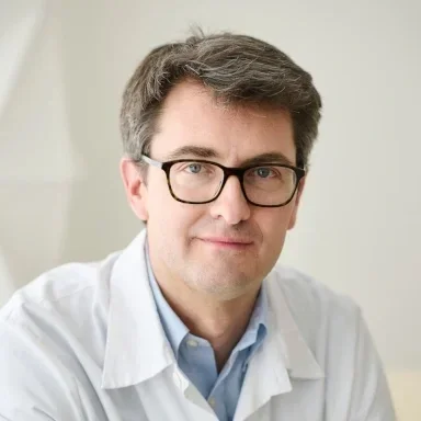 A man with dark hair, glasses, wearing a white lab coat, smiling in a medical or professional setting.