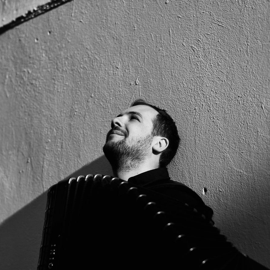 A black and white photograph of a man with a beard, smiling with eyes closed, leaning against a textured wall with shadows cast on it.