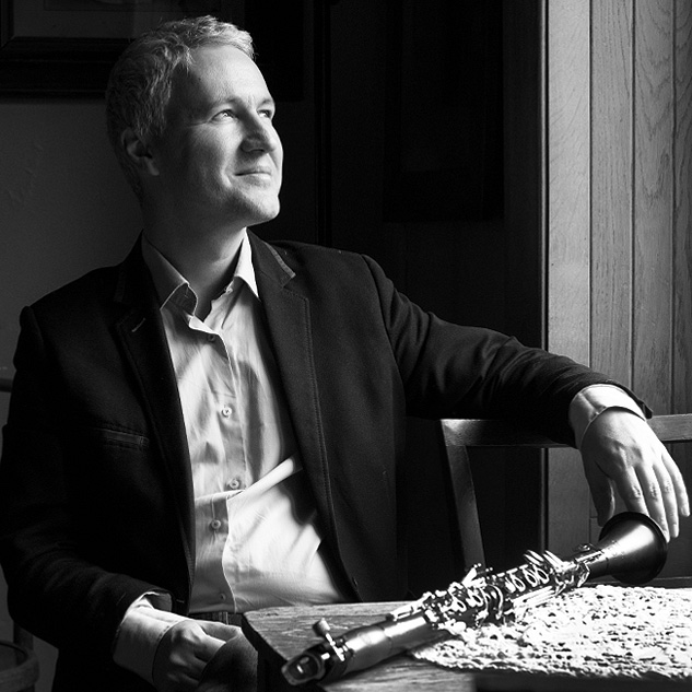 A man in a suit sitting at a wooden table with a telescope, looking out the window with a thoughtful expression in a black-and-white photo.