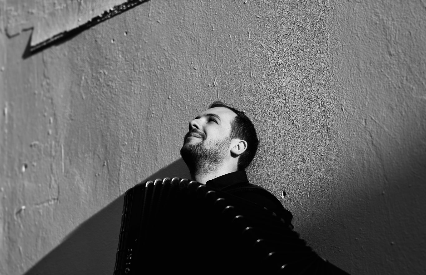 Black and white photo of a man with a beard smiling and looking up, leaning against a textured wall with sunlight casting a shadow behind him.