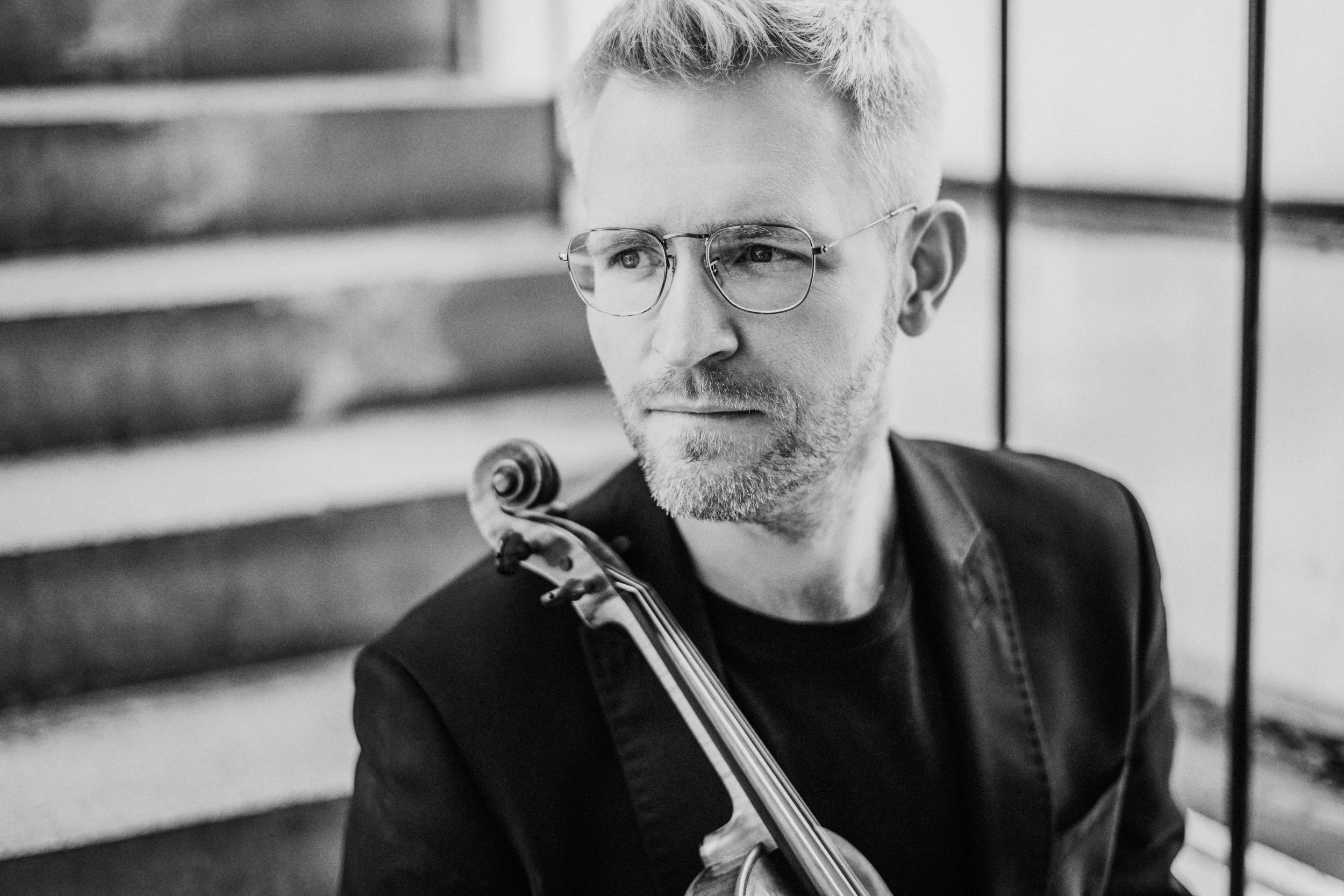 A man with glasses and a beard, holding a violin, sitting on a staircase, looking to the side.