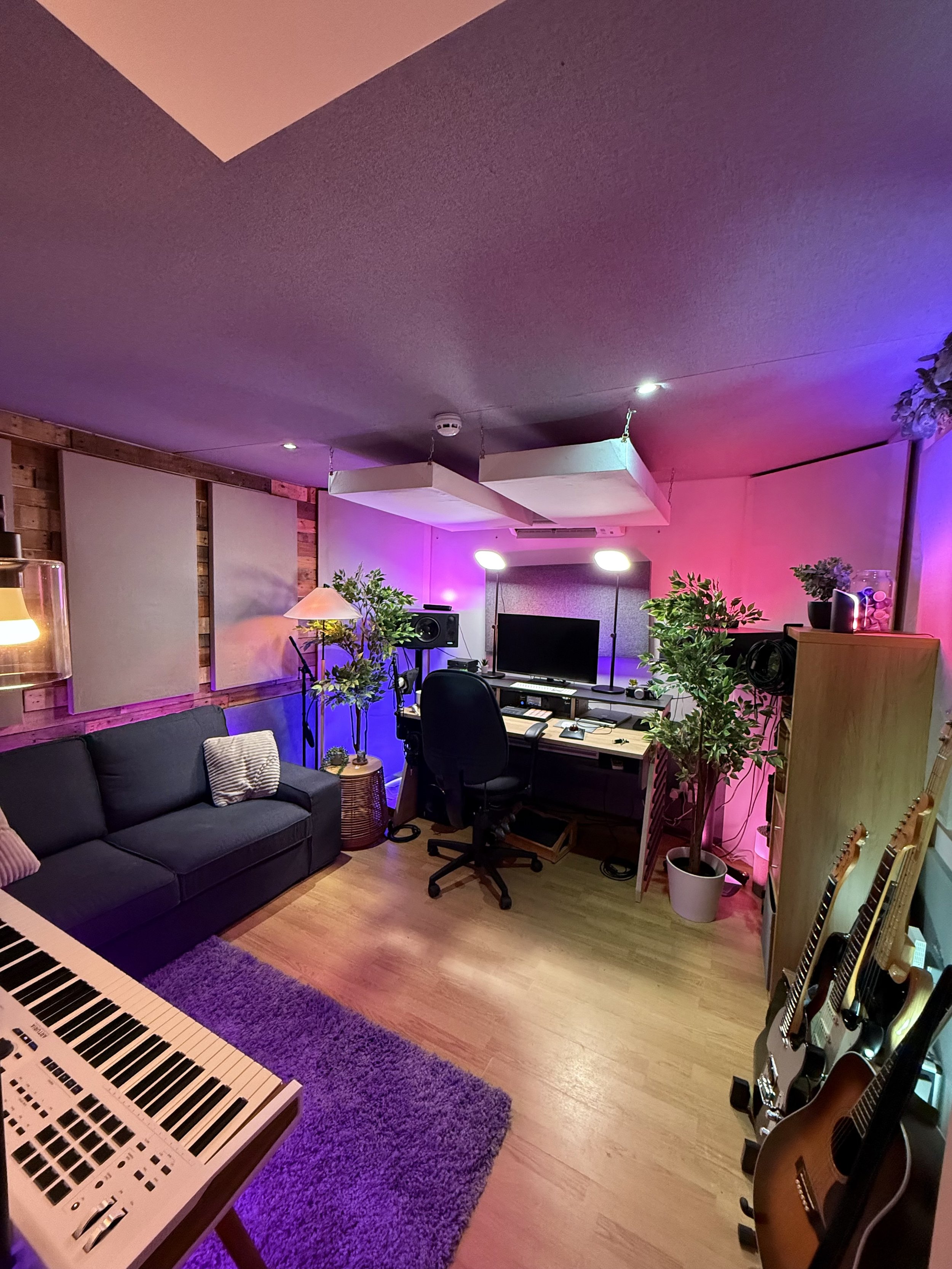 Music studio with a black sofa, a keyboard, a purple rug, potted plants, guitars, a TV, and recording equipment, illuminated with colorful purple and pink lighting.