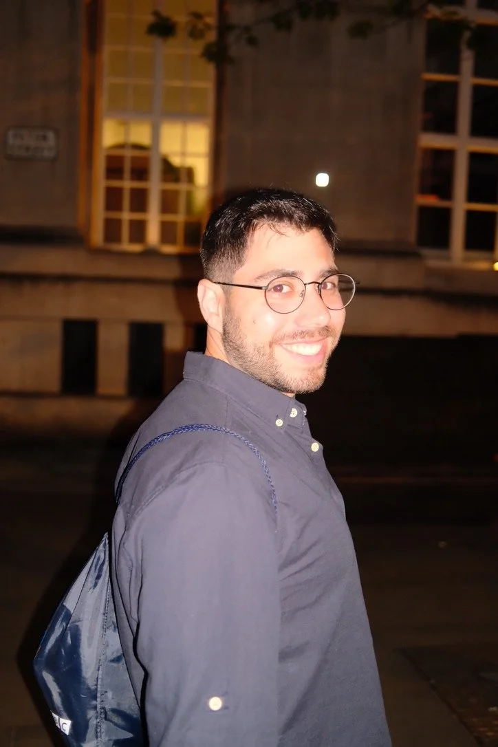 A young man with short dark hair, glasses, and facial hair, smiling at the camera, wearing a dark button-up shirt, standing outdoors at night with a building and illuminated windows in the background.