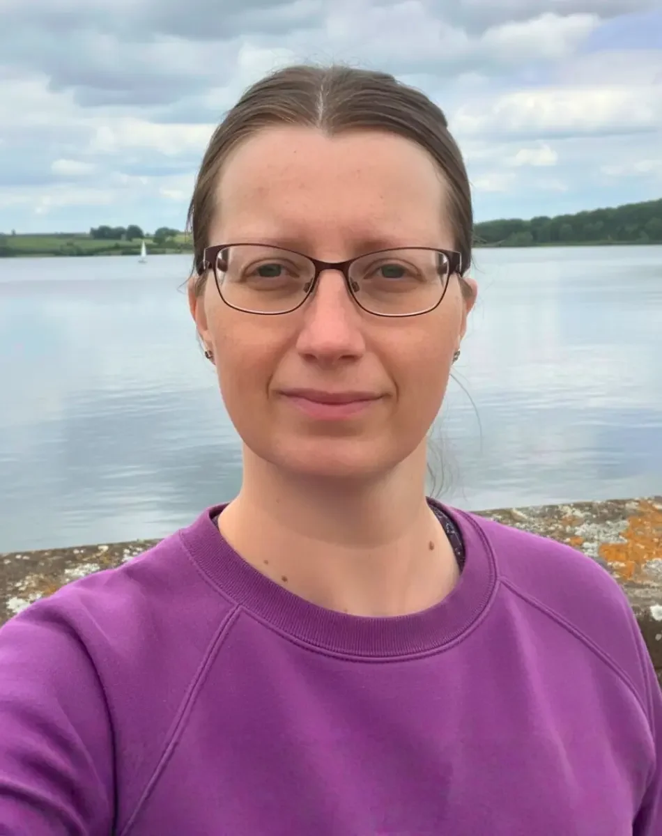 A woman with glasses and brown hair pulled back, wearing a purple sweatshirt, standing outdoors near a body of water with a cloudy sky behind.