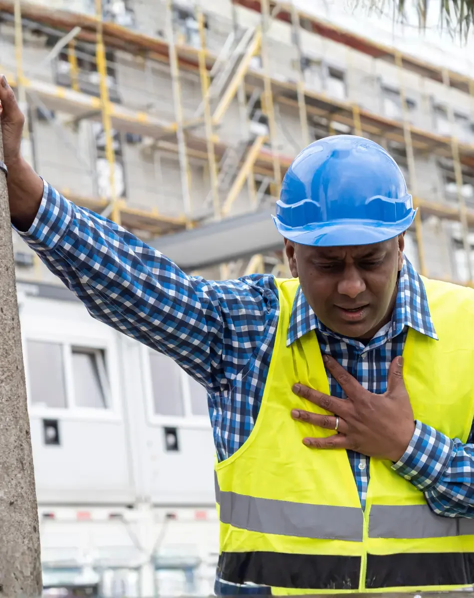 Construction worker in a blue hard hat and reflective vest clutching his chest with a pained expression, standing outdoors at a construction site.