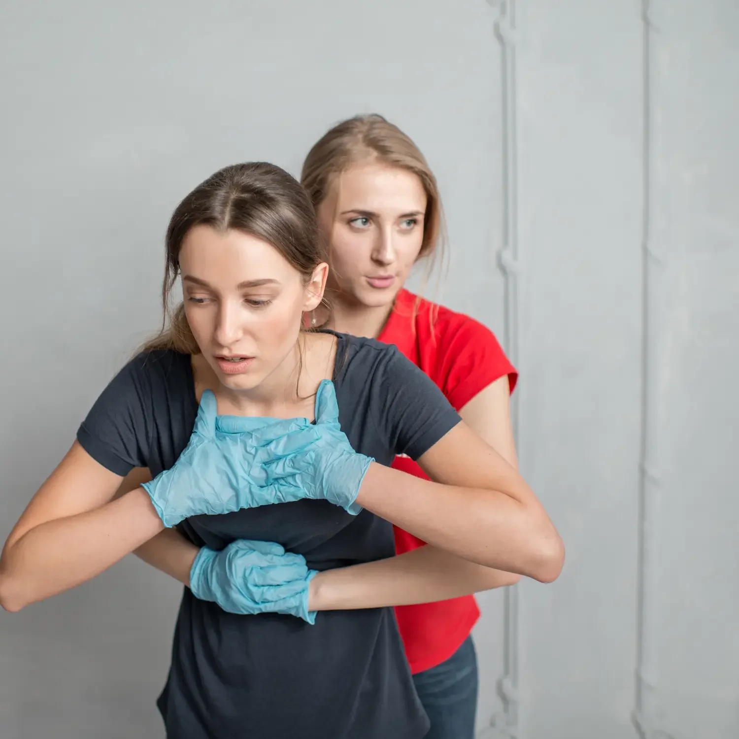 Two women, one in a black shirt and another in a red shirt, with the woman in black showing signs of difficulty, being assisted by the woman in red, who is holding her from behind in a supportive manner.