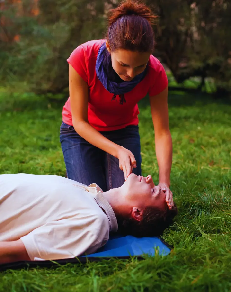 A woman performing CPR on a woman lying on the grass outdoors.