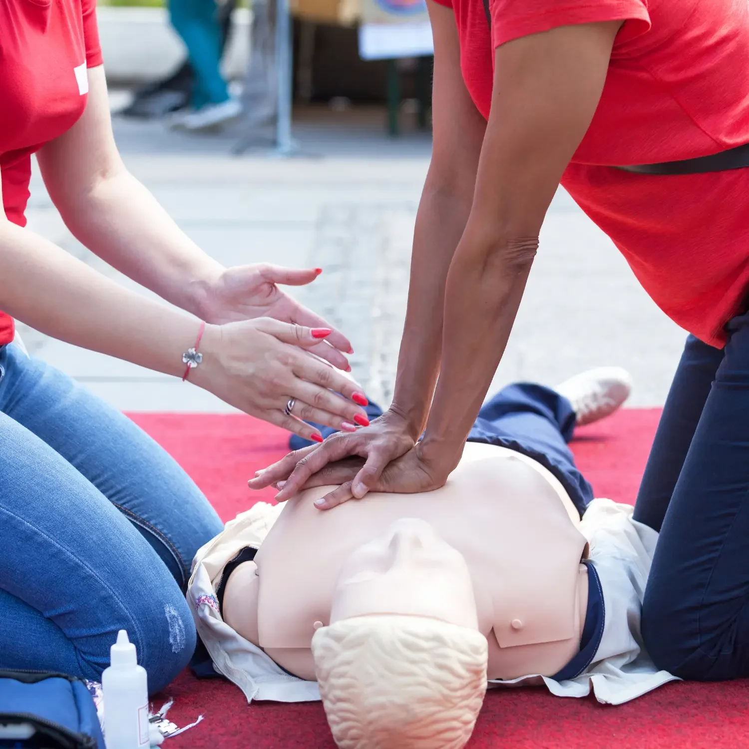 Two individuals practicing CPR on a medical training mannequin, with one performing chest compressions and the other preparing to assist, on a red carpet outdoors.