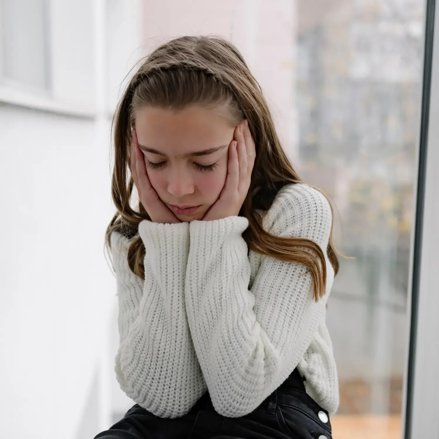 A girl with long brown hair and closed eyes, wearing a white knitted sweater, sitting near a large window with an outdoor view, holding her head with both hands, appearing distressed or upset.