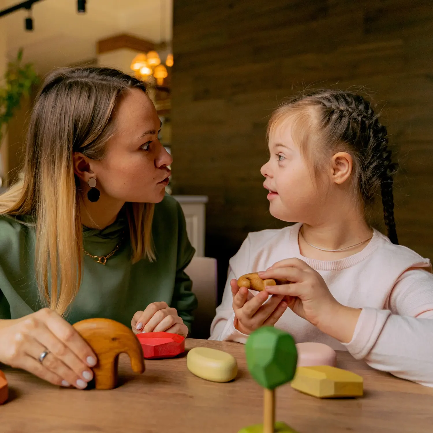 A woman and a young girl face each other across a table with colorful wooden toys, engaging in a conversation or playing.