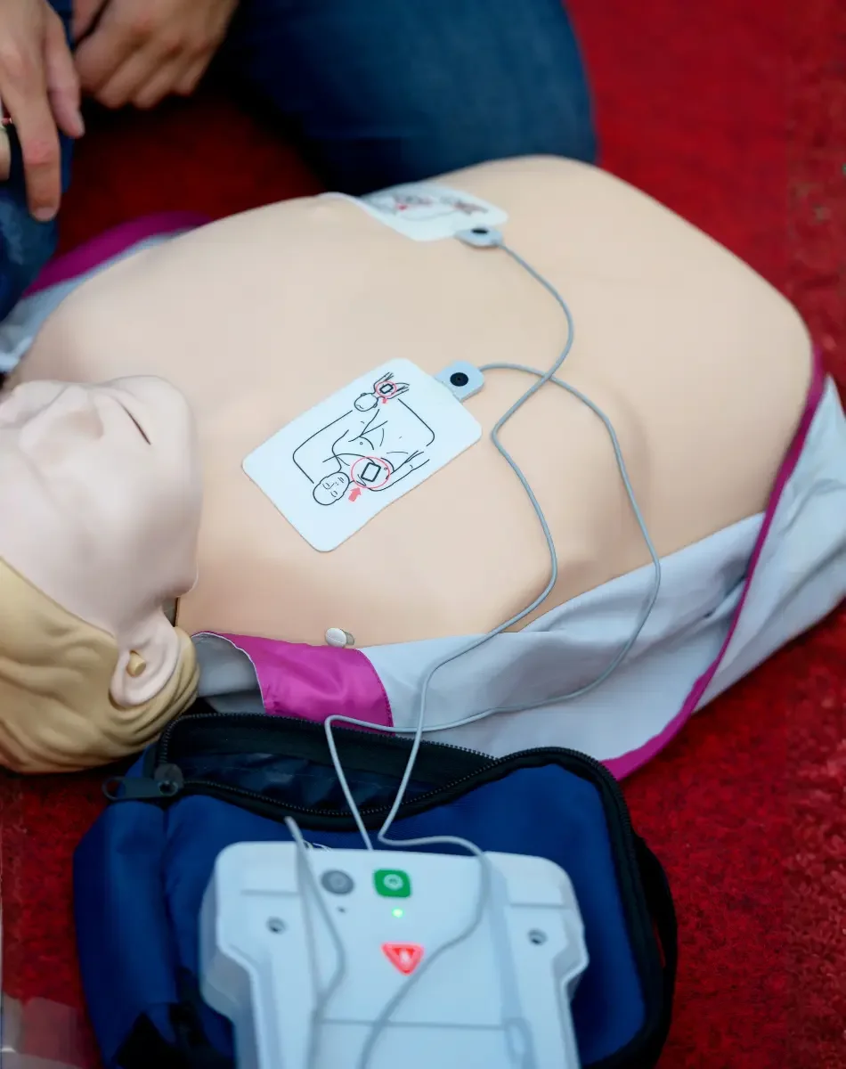 A mannequin lying on the floor with a CPR manikin on its chest, connected to a defibrillator via wires, used for CPR training.