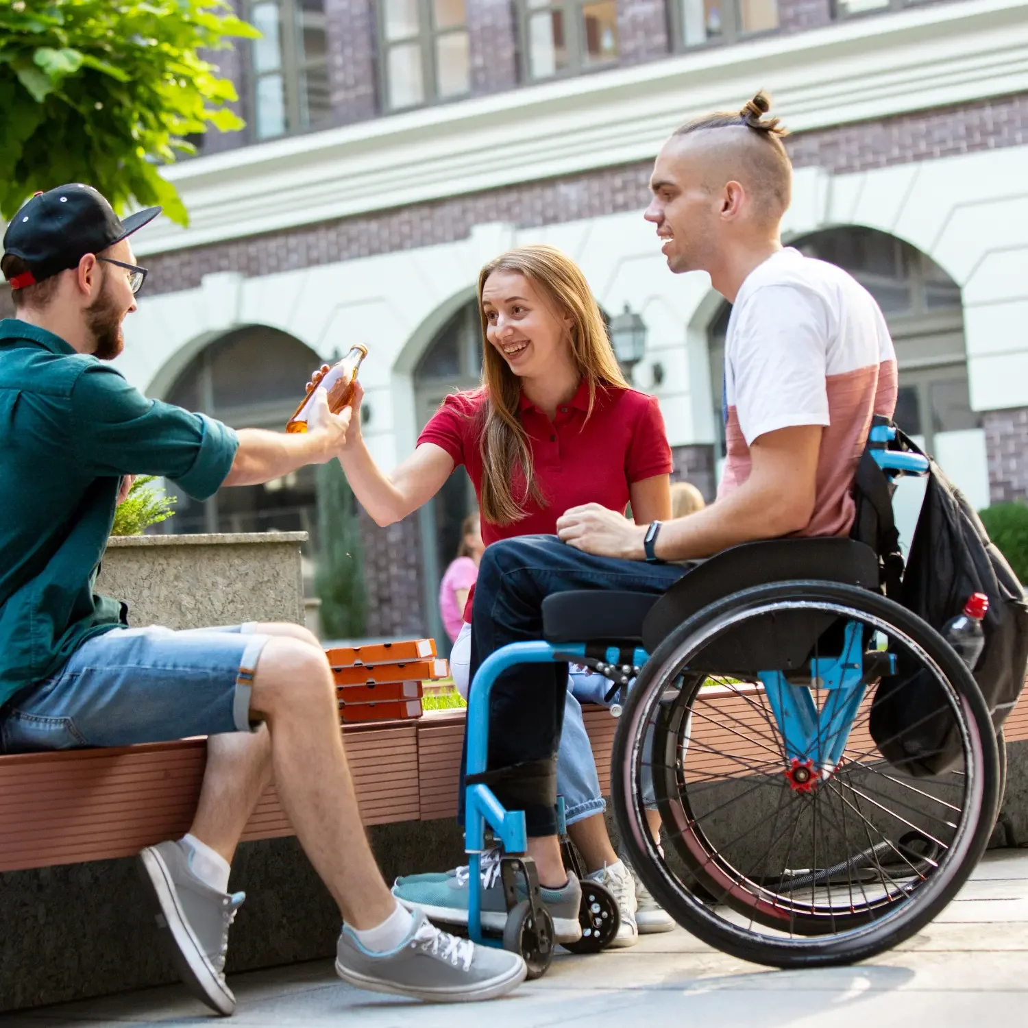 A young woman in a red shirt and man in a wheelchair sharing a drink with a young man in casual clothes on a city street during daytime.
