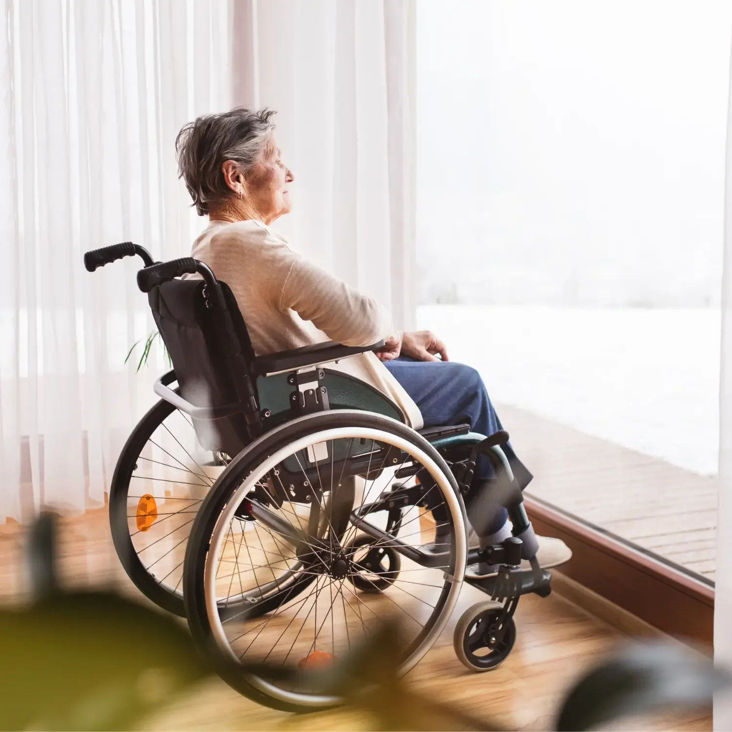 An elderly woman with gray hair sitting in a wheelchair, looking out a large window with sheer curtains.