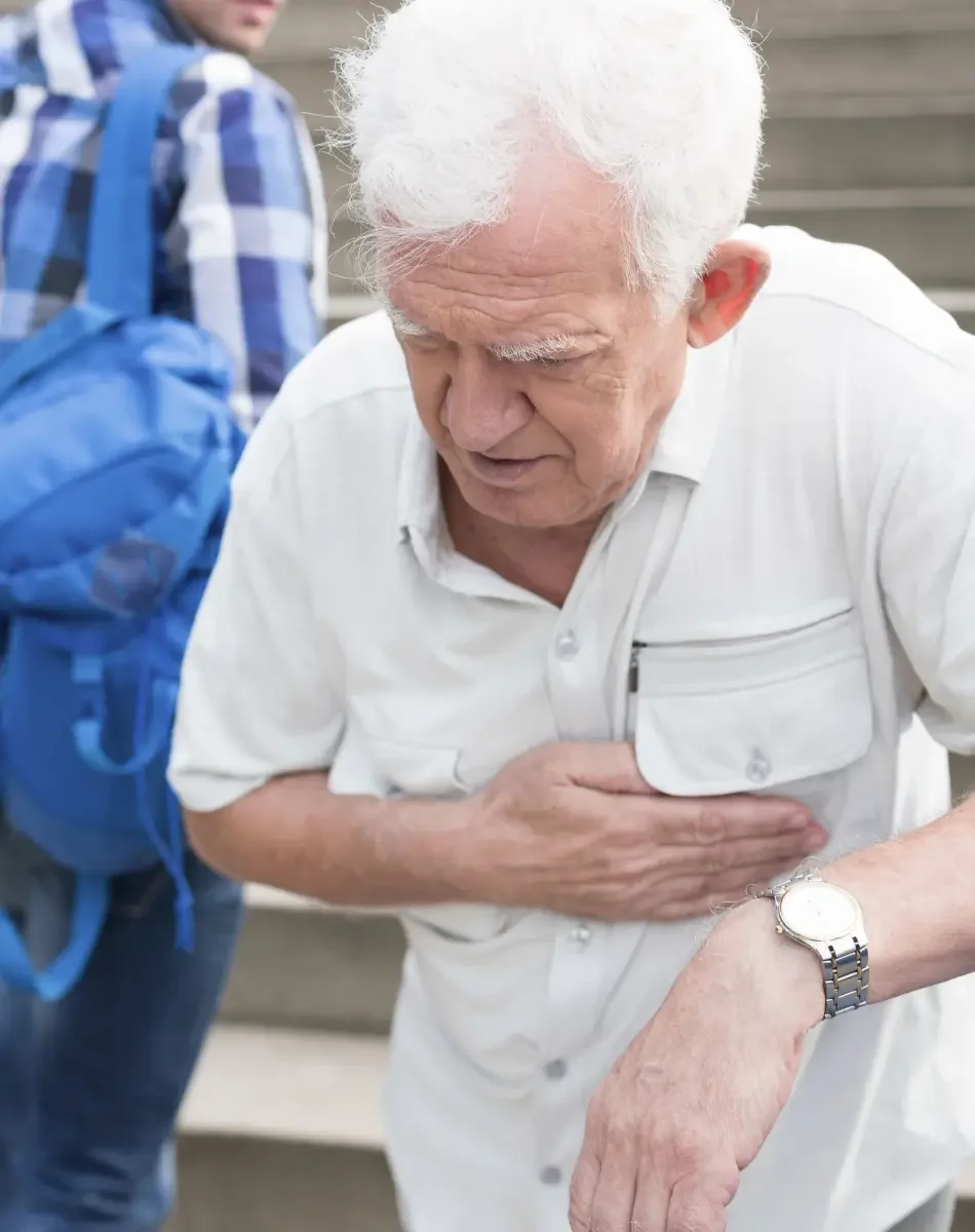 An elderly man with white hair and a white shirt is clutching his chest, appearing distressed or experiencing chest pain. A person with a blue checkered shirt and a blue backpack stands nearby in the background.