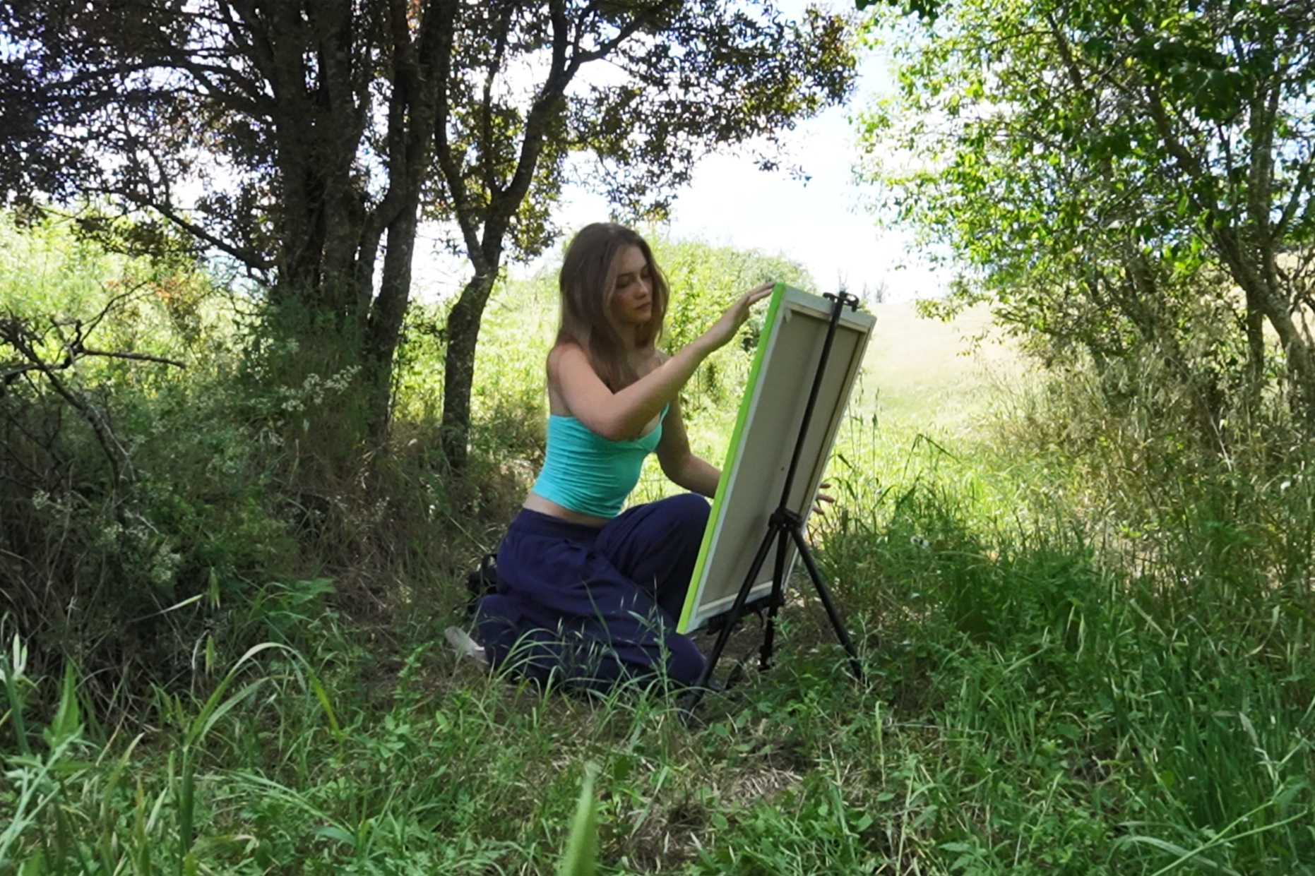 A woman sitting outdoors in a grassy area surrounded by trees, painting on an easel.