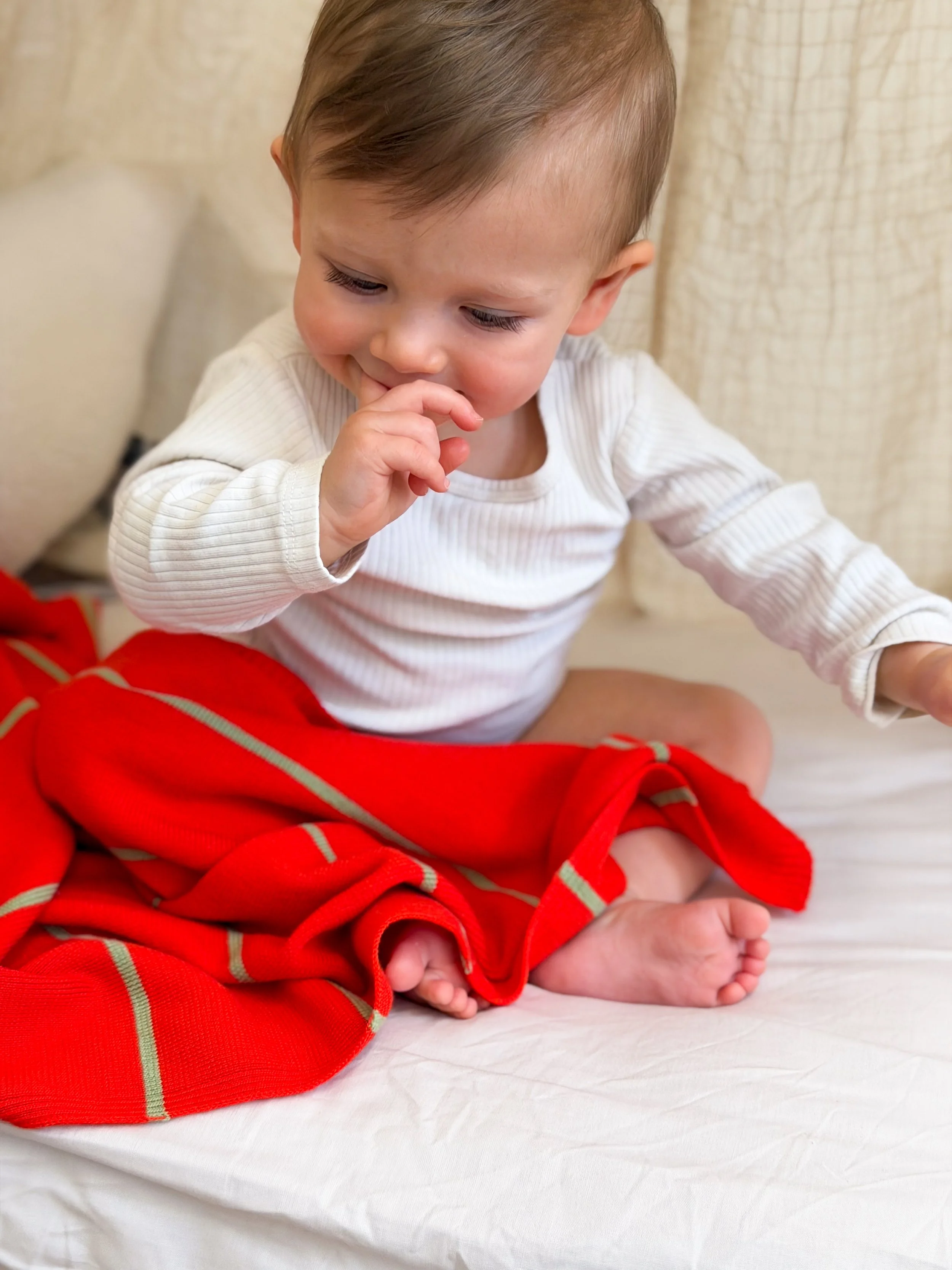 Baby sitting on the floor with a white body and a colourfull Mellrose merino blanket in bloodaronge.