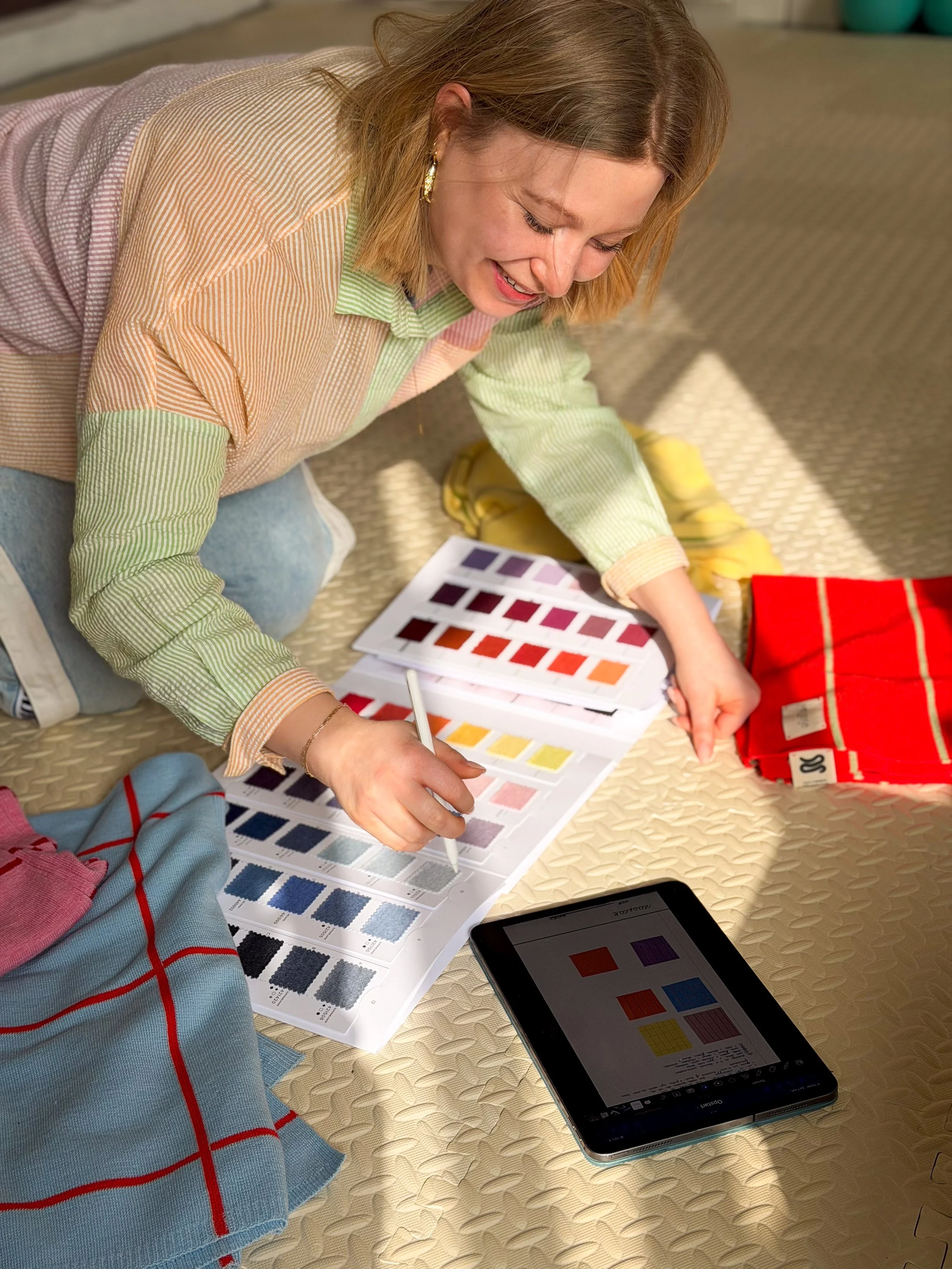 A woman is sitting on the floor, holding a color swatch fan deck and a pen, looking at color swatches, with a tablet displaying color options nearby. She is surrounded by clothing fabrics and has a smile on her face.