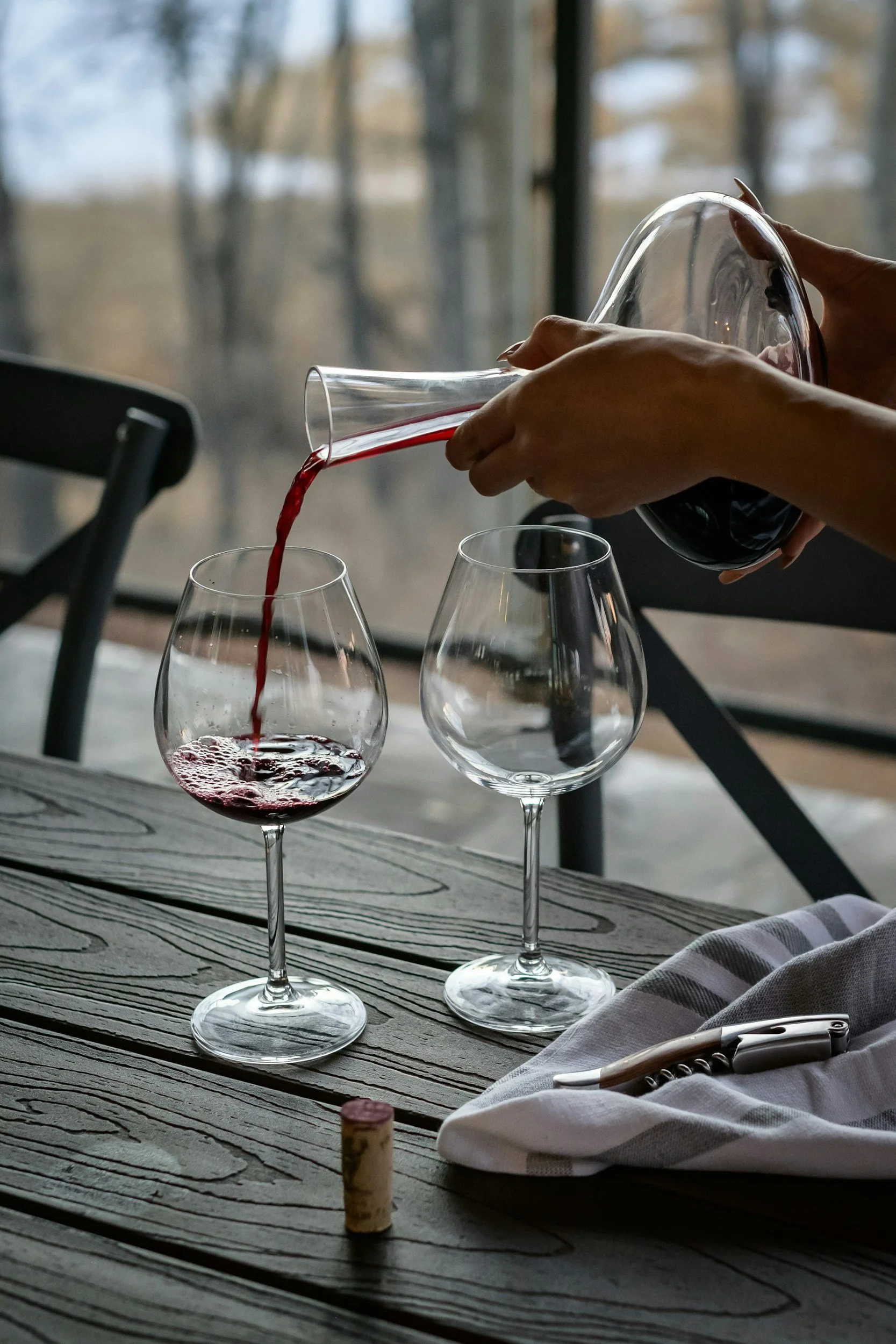 A person pouring red wine from a decanter into a glass on a rustic wooden table. There's an empty wine glass next to it, a cork, a napkin, and a corkscrew. The setting appears to be indoors near a window with a blurred outdoor view.