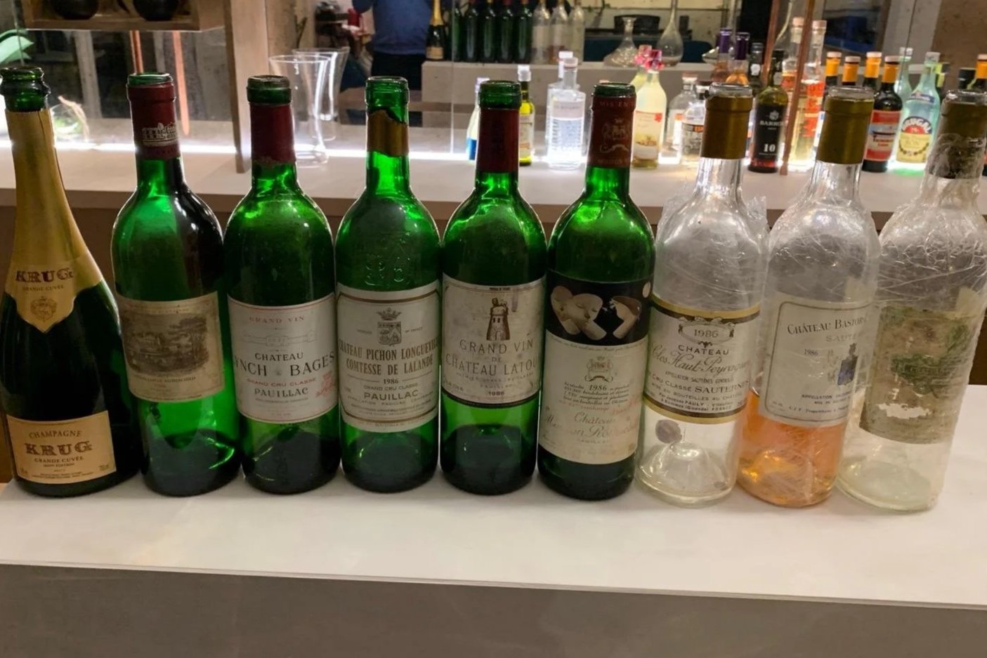 Assorted bottles of wine and champagne displayed on a white surface in a bar or restaurant setting, with a mirror and various liquor bottles in the background.