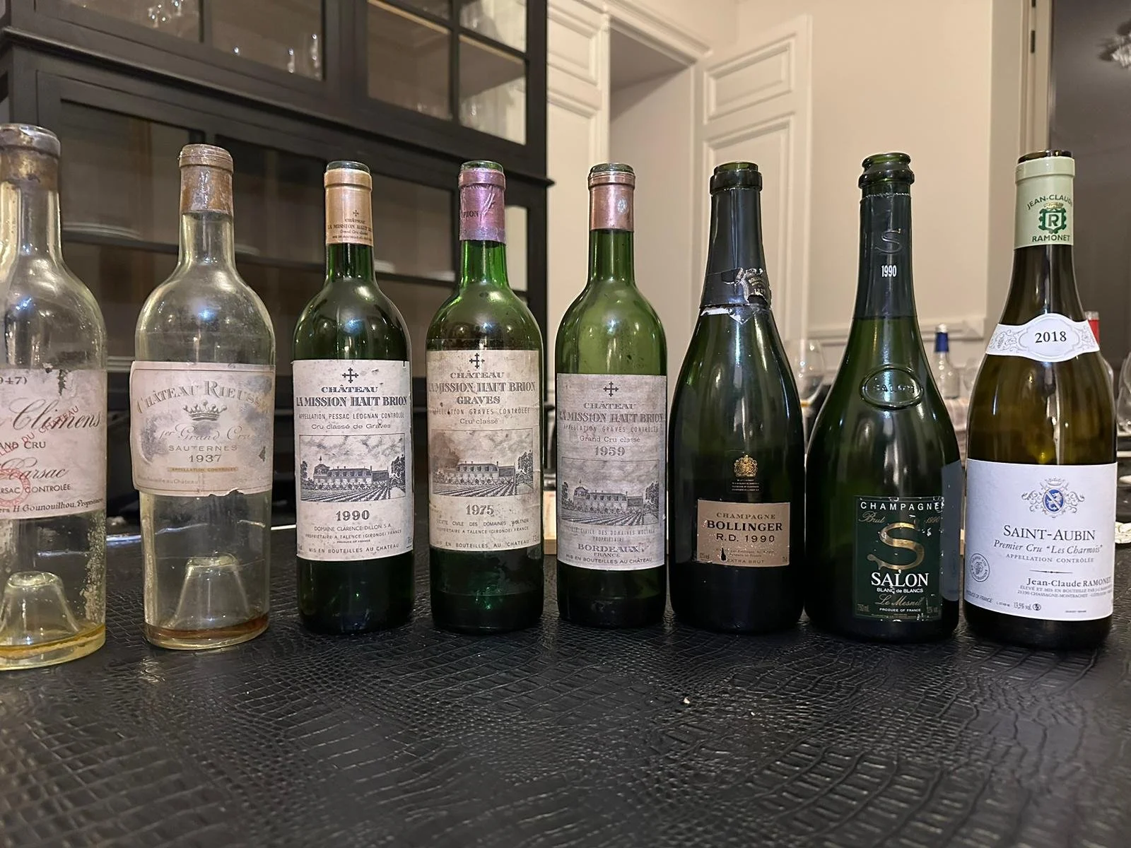 A row of eight empty and partially filled wine bottles from different years and types, lined up on a table in a room with white cabinetry and a textured black surface.