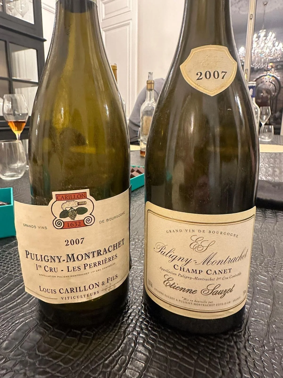 Two bottles of Puligny-Montrachet wine from 2007 on a textured black table, with glasses and other bottles in the background.