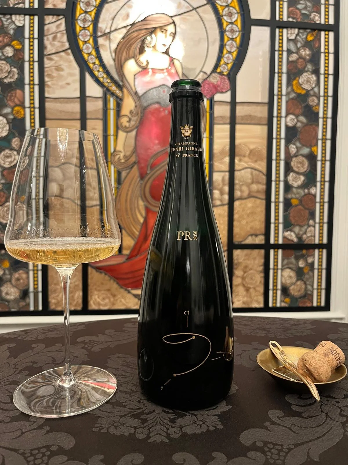 A glass of white wine, a black bottle of champagne, and a small gold dish with a corkscrew and cork are on a dark tablecloth with a floral pattern. In the background, there is a stained glass window depicting a woman with long flowing hair in a reddi