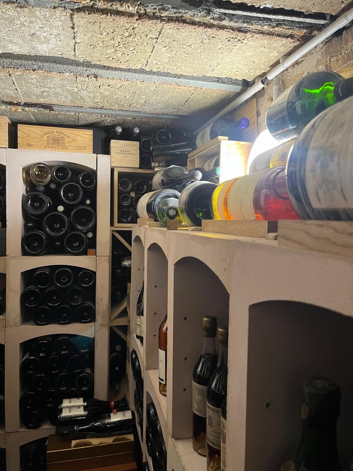 A cellar or wine storage room with wooden wine racks filled with bottles of wine, some stored horizontally and some horizontally on shelves, and a concrete ceiling with pipes.