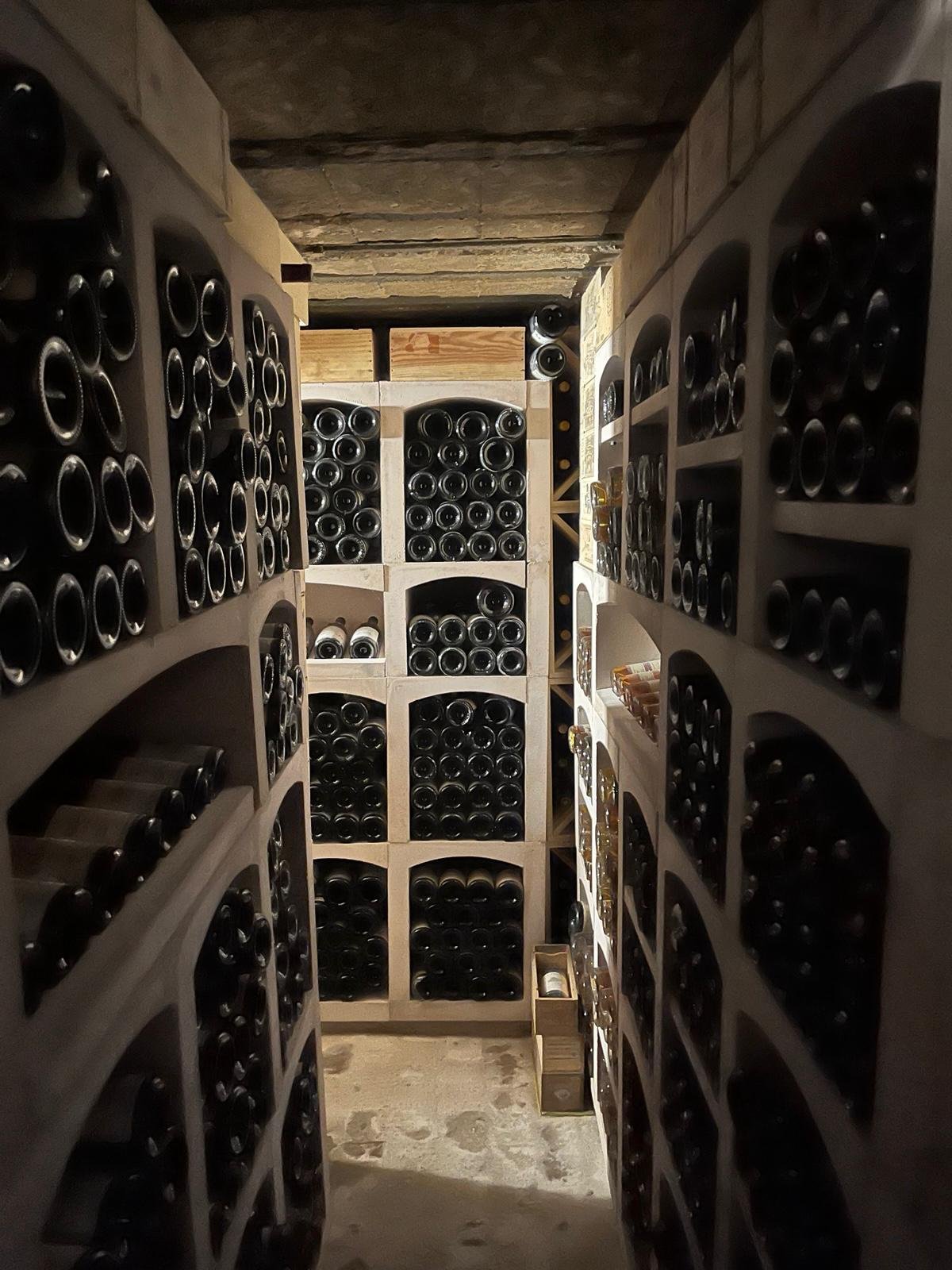 A wine cellar with beige wooden wine racks filled with bottles of wine, lit by dim lighting.