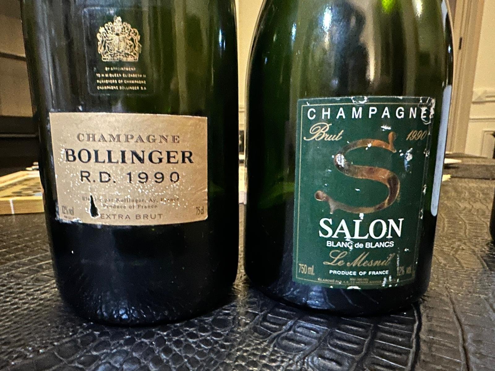 Two vintage champagne bottles. The left bottle has a beige label reading "Champagne Bollinger R.D. 1990 Extra Brut." The right bottle has a green label with gold and white text, reading "Champagne Brut Salon Blanc de Blancs Le Mesnil" from France.