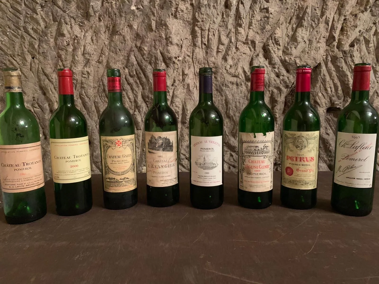 A row of seven vintage Bordeaux wine bottles with various labels, some with red wax seals, displayed on a wooden surface against a stone wall background.
