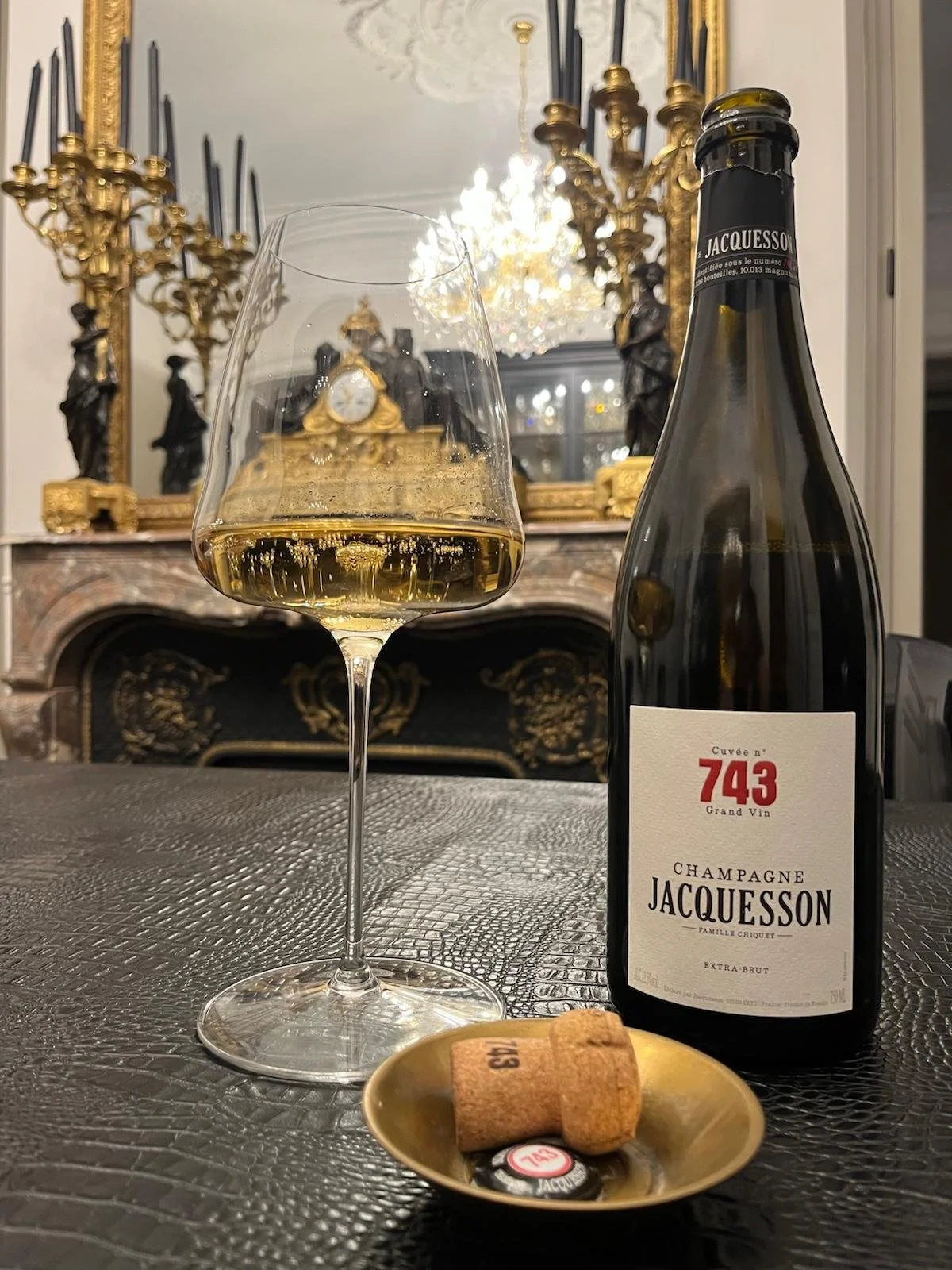 A glass of white wine, a bottle of Jacquesson champagne, and a cork with a seal on a textured black table, with a luxurious ornate fireplace and mirror decorated with gold accents and statuettes in the background.