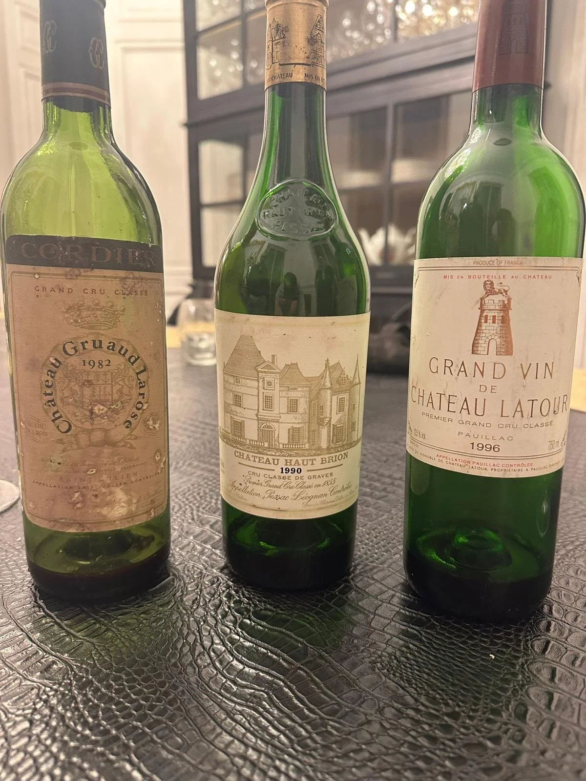 Three vintage wine bottles on a textured table, including Château Gruaud Larose 1982, Château Haut Brion 1990, and Château Latour 1996.