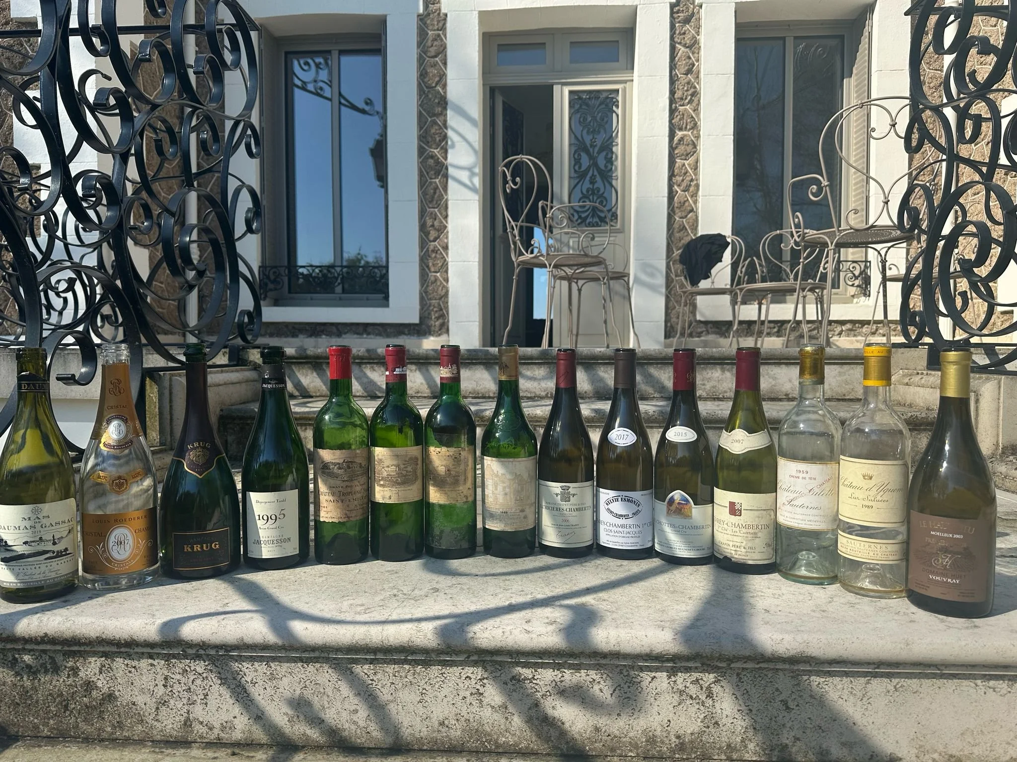 A row of empty wine bottles lined up on a concrete surface outside a house with stairs and outdoor furniture in the background.