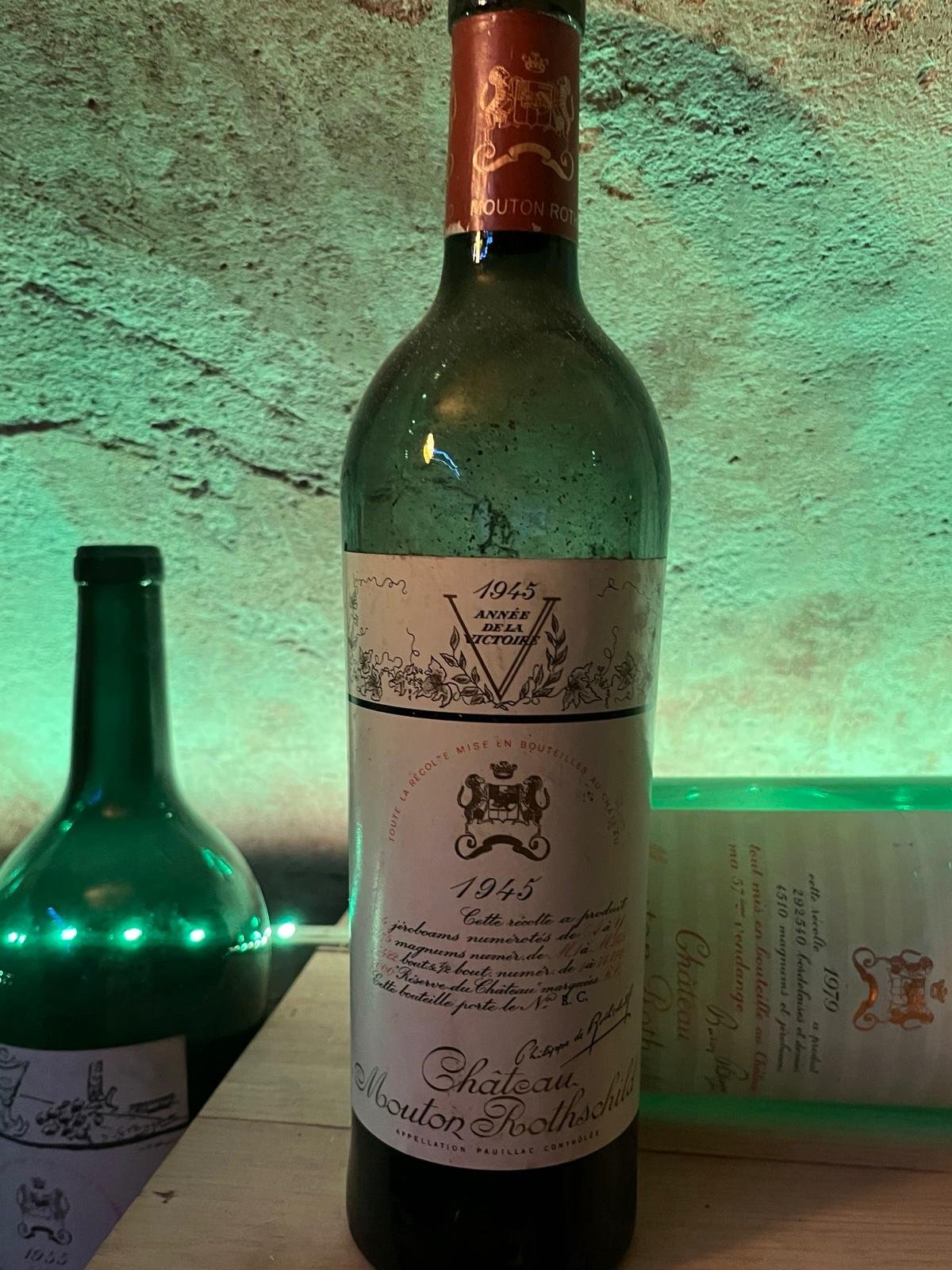 A 1945 bottle of Château Mouton Rothschild wine displayed on a surface, with a decorative background and another green bottle partially visible beside it.