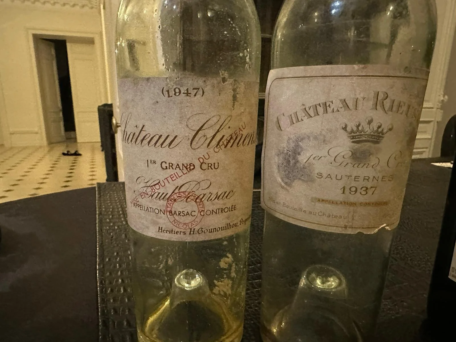 Two vintage glass wine bottles with faded labels featuring Château Chateau de Barsac from 1947 and 1937, placed on a dark surface in an elegant room with a patterned floor and white paneling in the background.