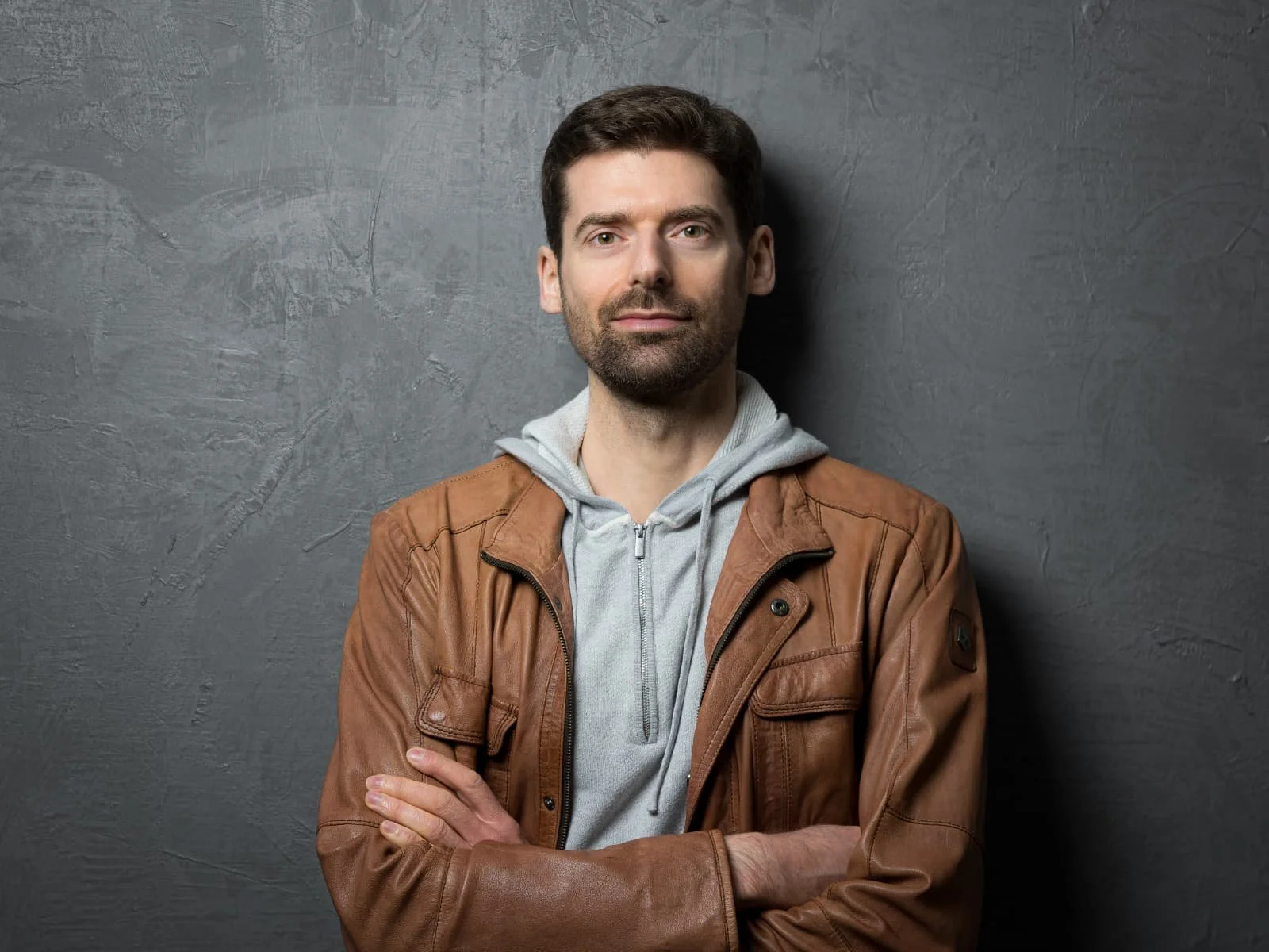 Alessandro Limentani. A man with dark hair and beard, arms crossed, wearing a brown leather jacket over a gray hoodie, standing against a textured gray wall.