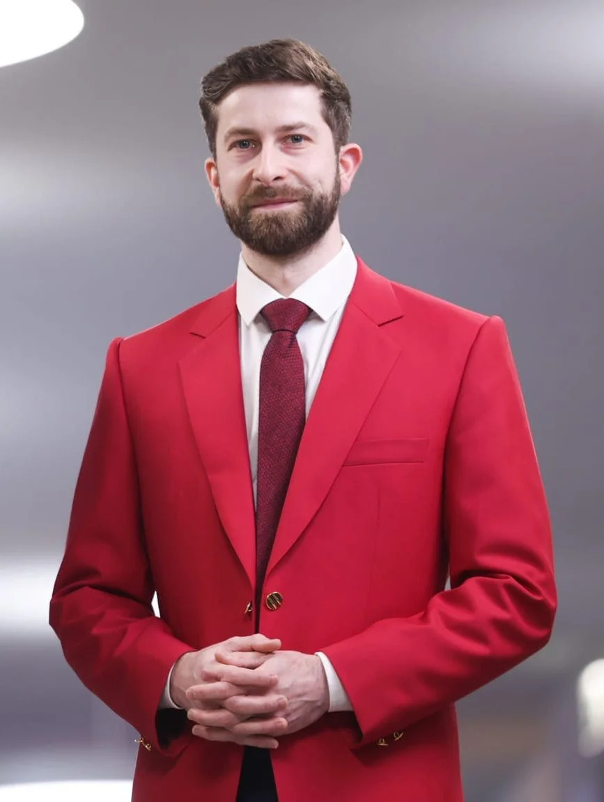 Christoph Weiß. A man with brown hair and a beard wearing a red suit, white shirt, and red tie, standing in front of a blurred gray background.