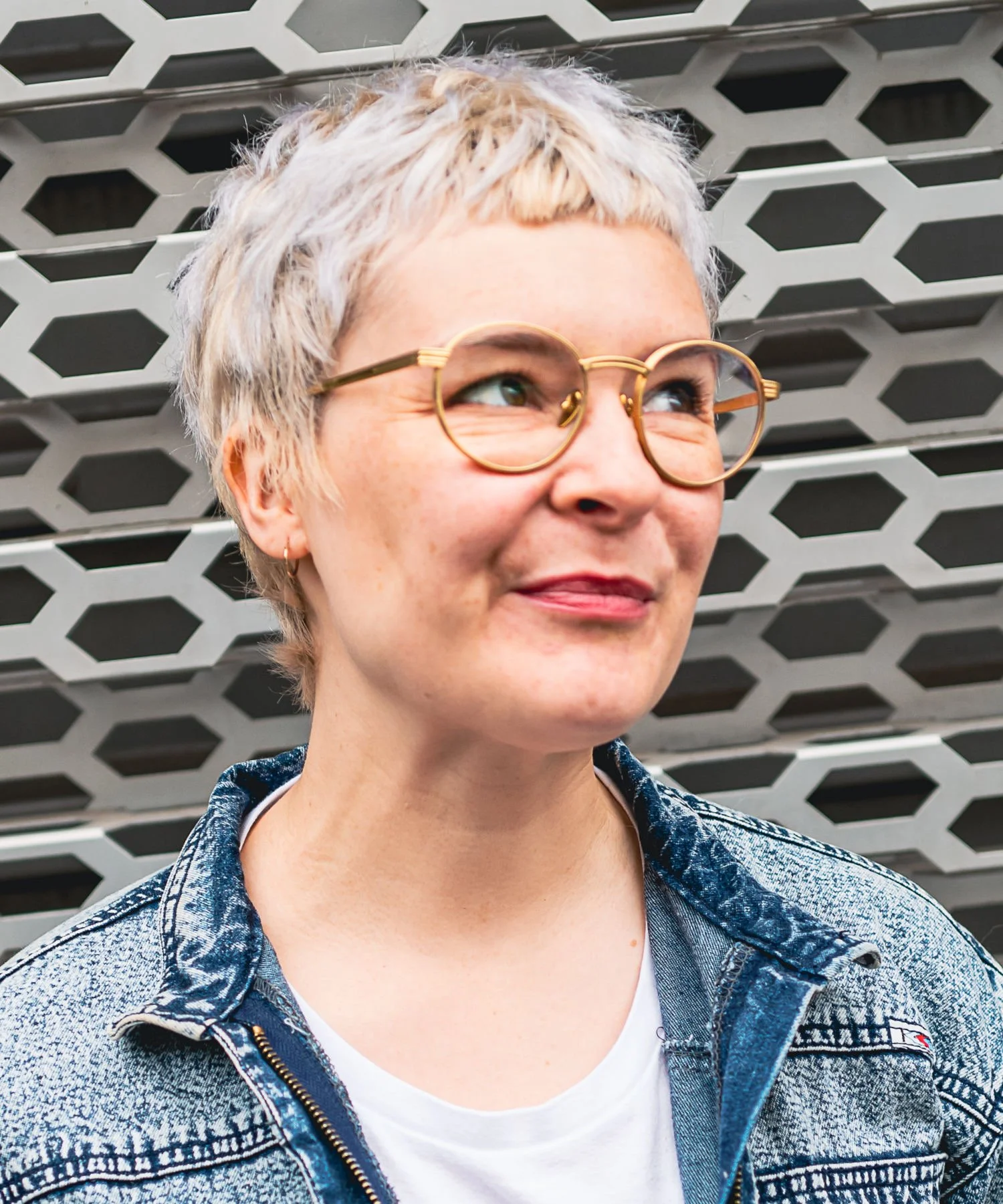 Anna-Sophia Lumpe. A woman with short, blonde, curly hair and glasses standing in front of a honeycomb-patterned metal wall.