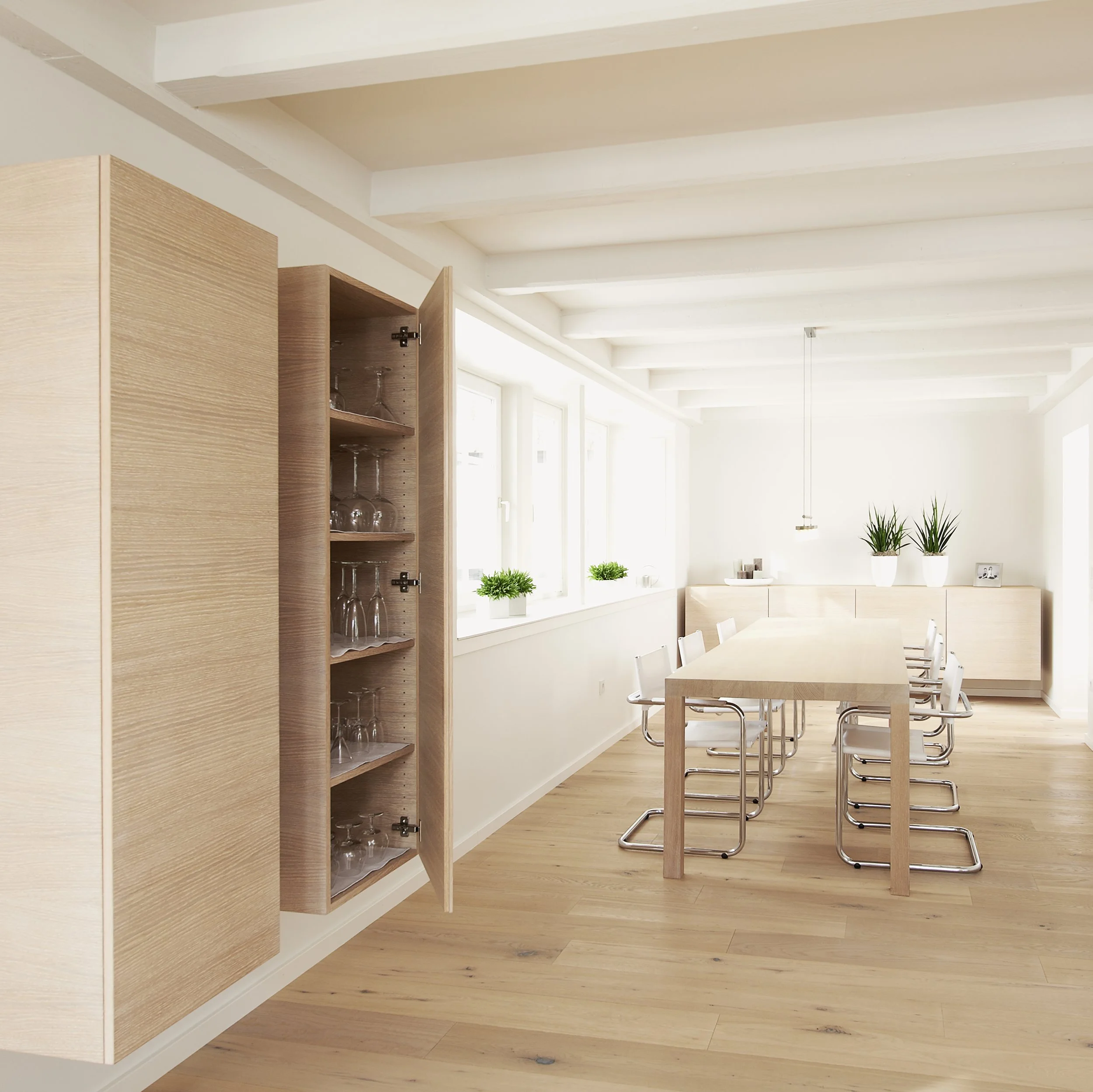 Heller Esszimmerraum mit Holztisch, weißen Stühlen, großen Fenstern, Topfpflanzen und einem weißen Sideboard.