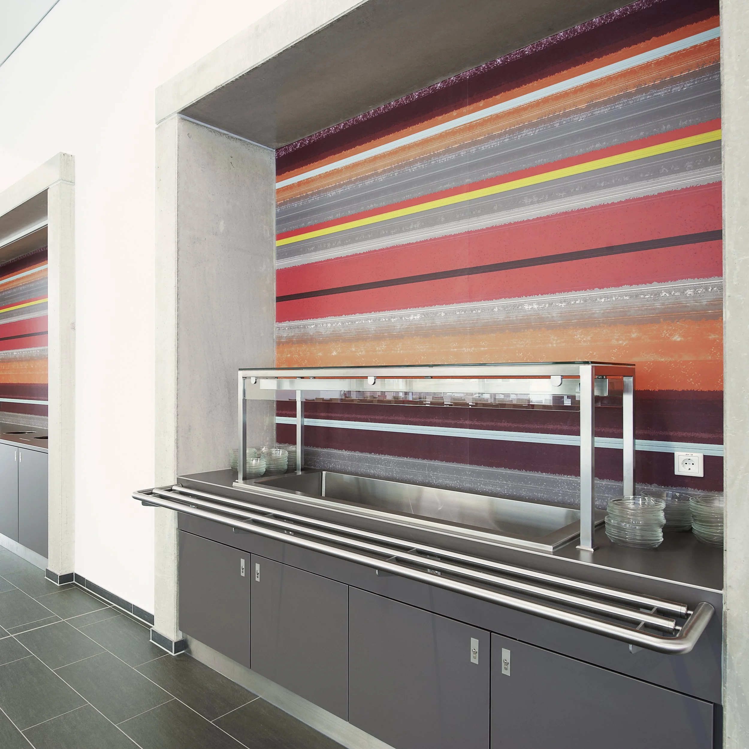 Kühlschrank mit einer Abdeckung, die in einer modernen Küche aufgestellt ist, vor einer farbenfrohen Wand mit horizontalen Streifen in Rot, Orange, Grau und Gelb.